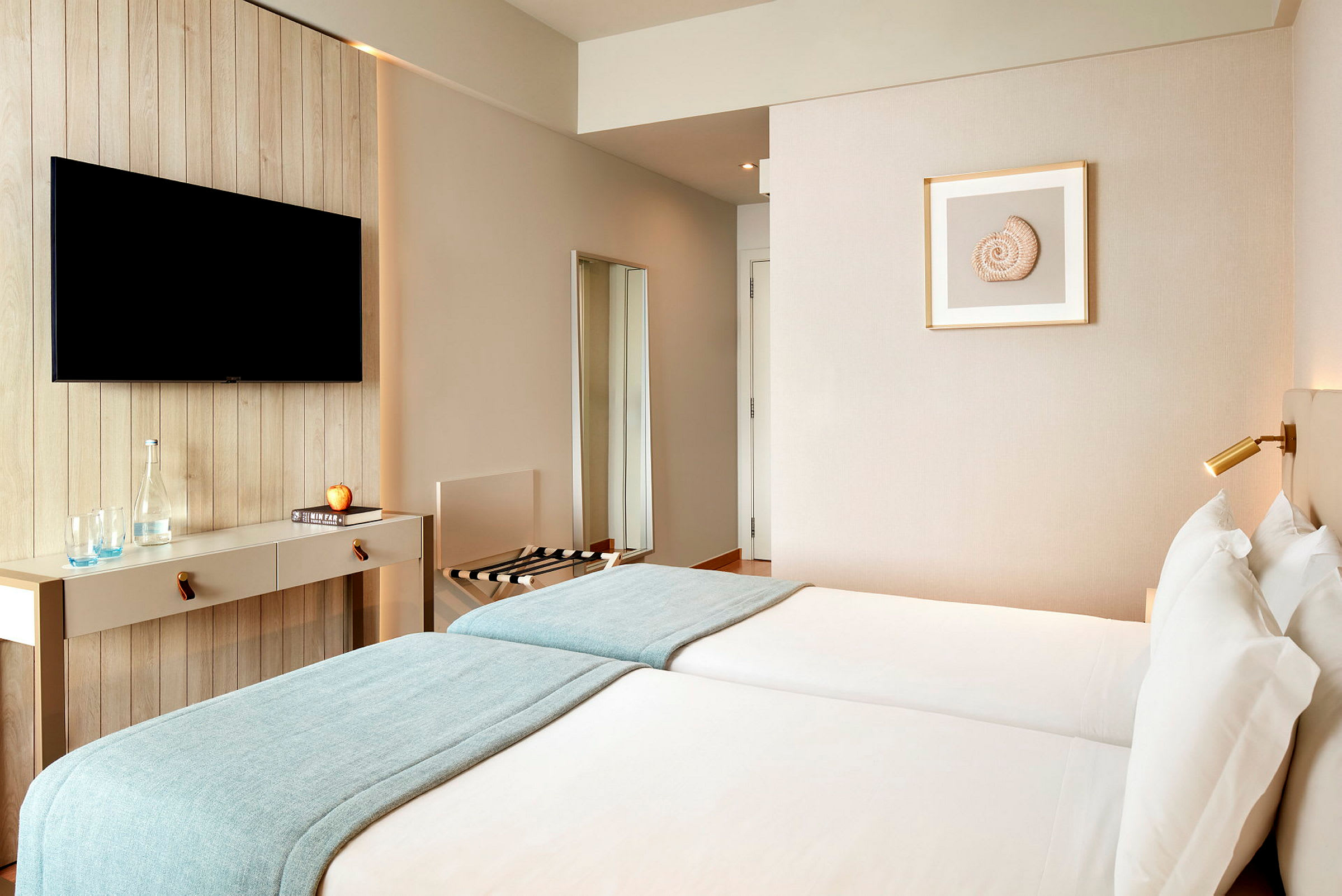 Et hotelværelse med to enkeltsenge, et vægmonteret TV og minimalistisk indretning. (AI-genereret tekst)