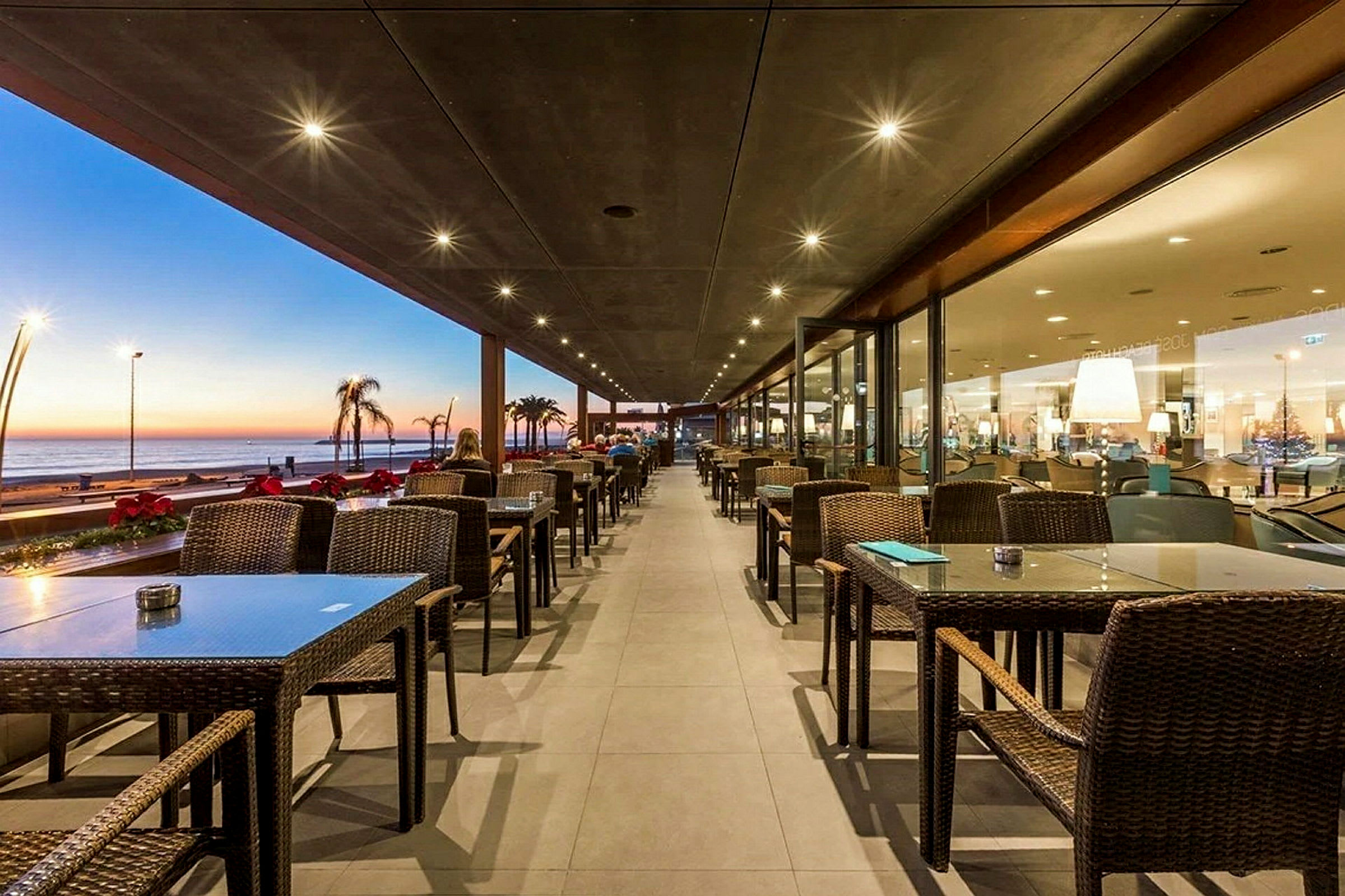Udendørsrestaurant med opstillet borde og stole, samt udsigt over havet under solnedgang. (AI-genereret tekst)