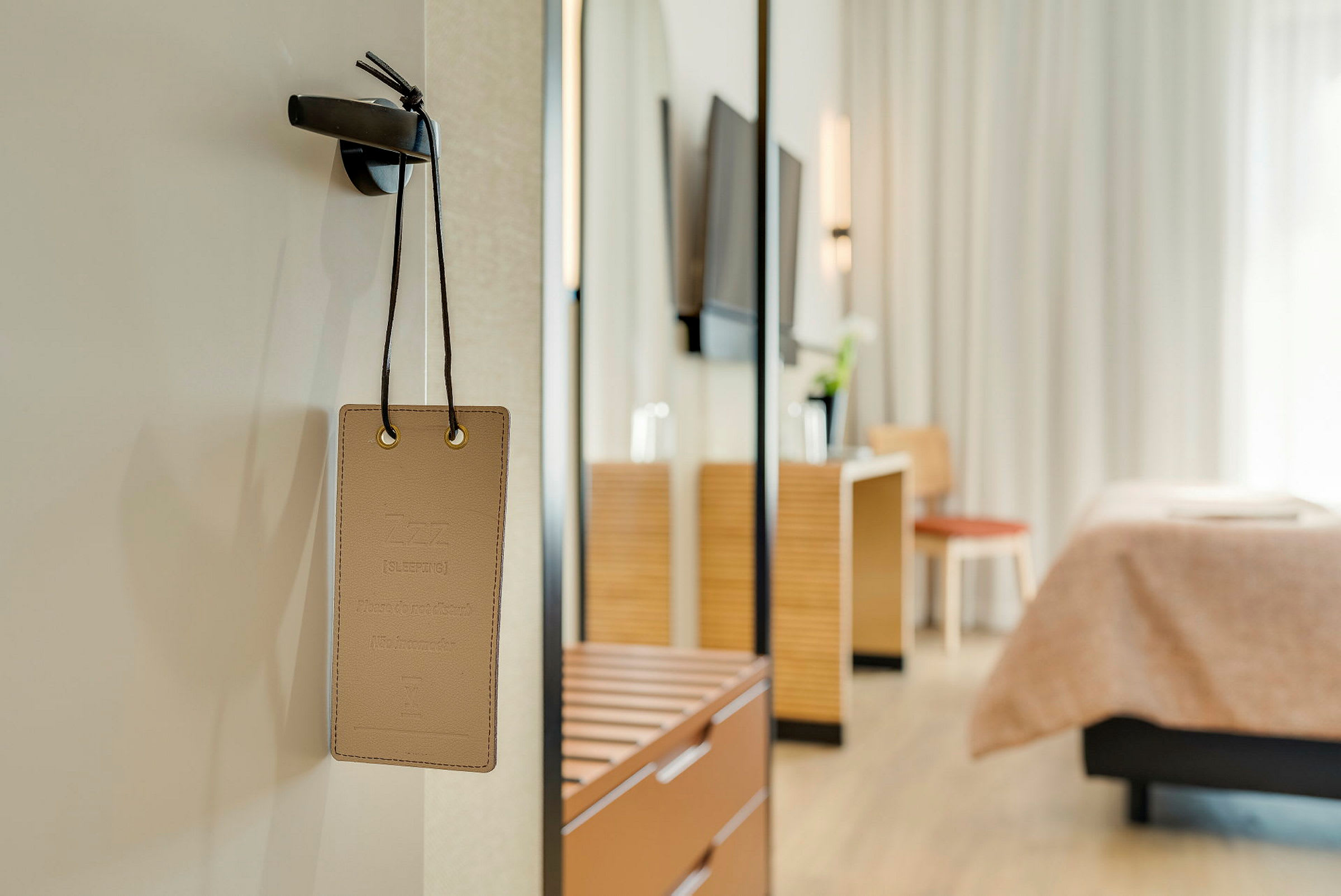 Skilt med teksten 'Sover, forstyr ikke' hængende på en dør med en hotelværelsesindretning i baggrunden. (AI-genereret tekst)