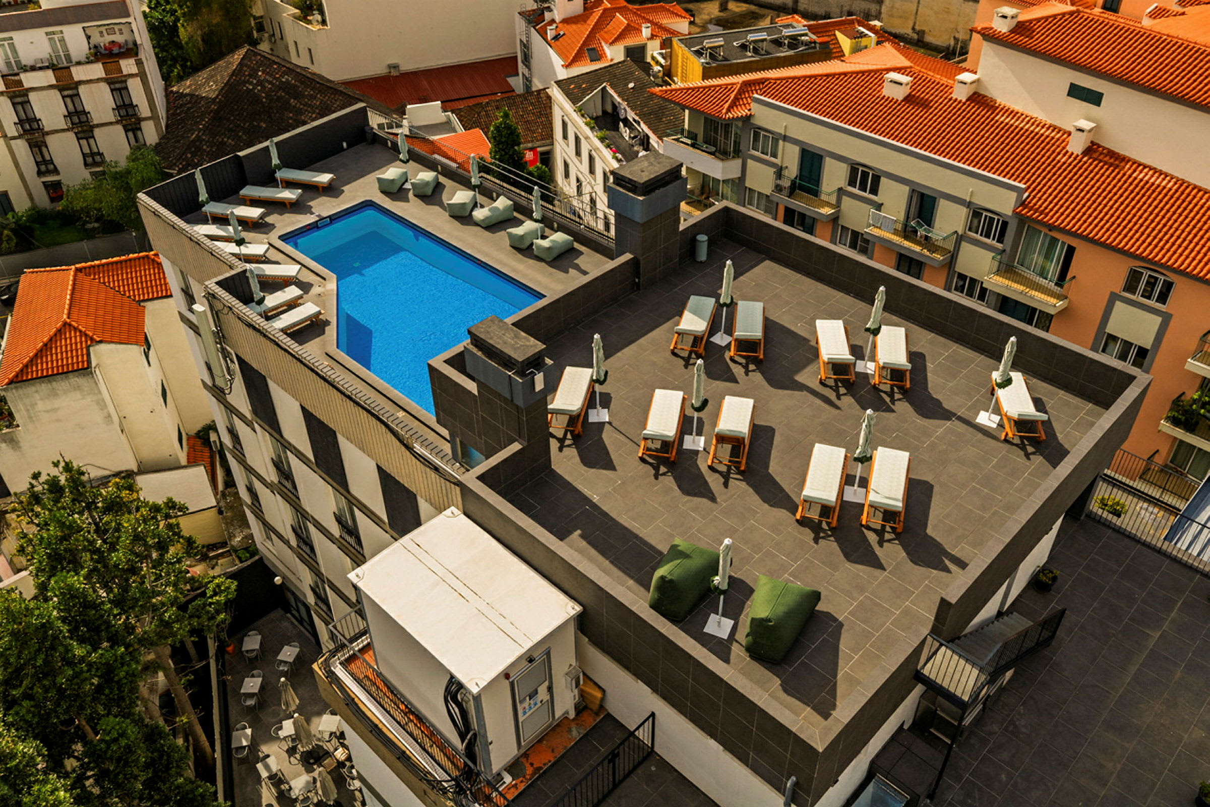 Billede af en tagterrasse med en blå pool, solsenge og parasoller, omgivet af bygninger med tegltage. (AI-genereret tekst)