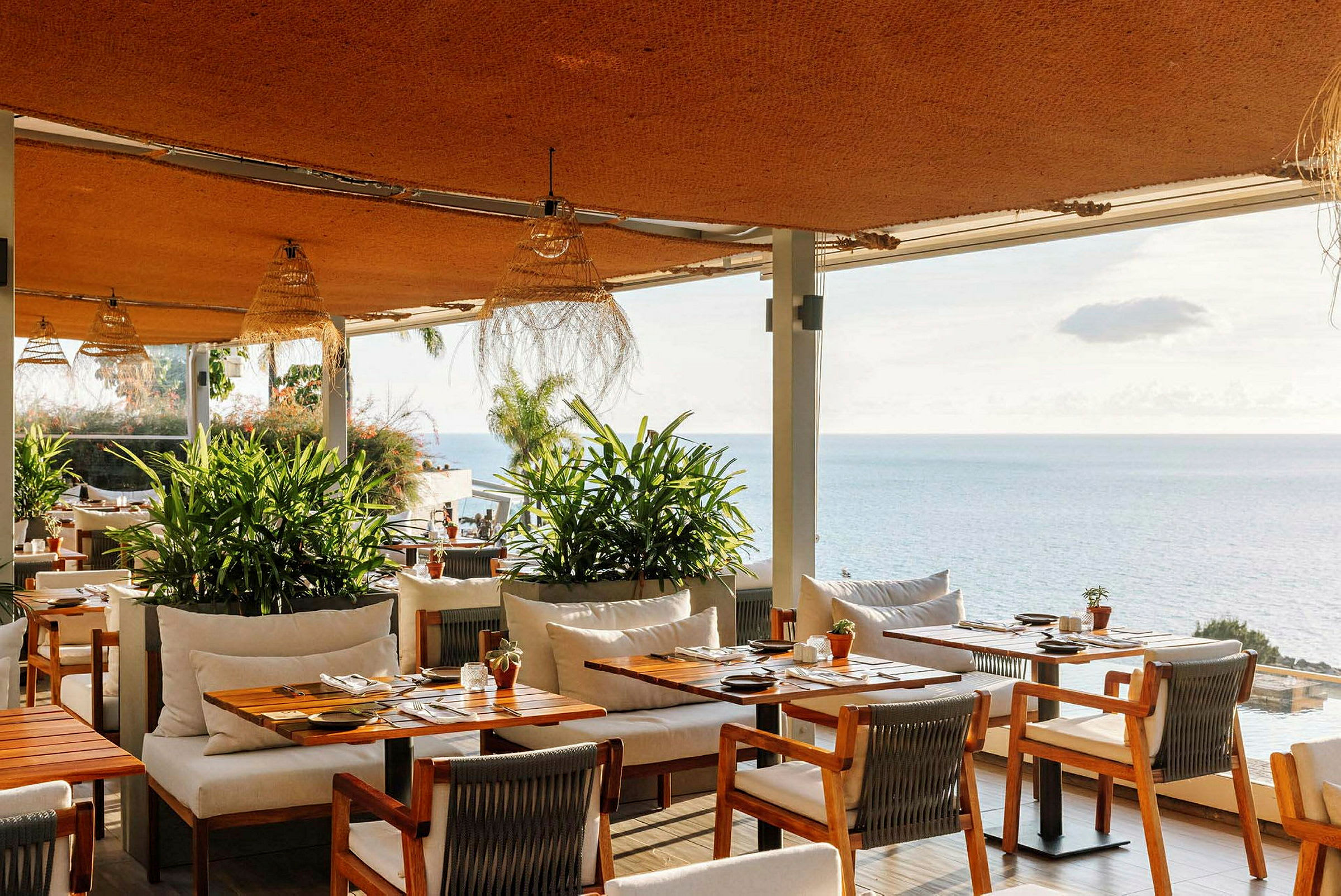 Udsigt over en restaurant med træborde og stole, grønne planter og havet i baggrunden. (AI-genereret tekst)