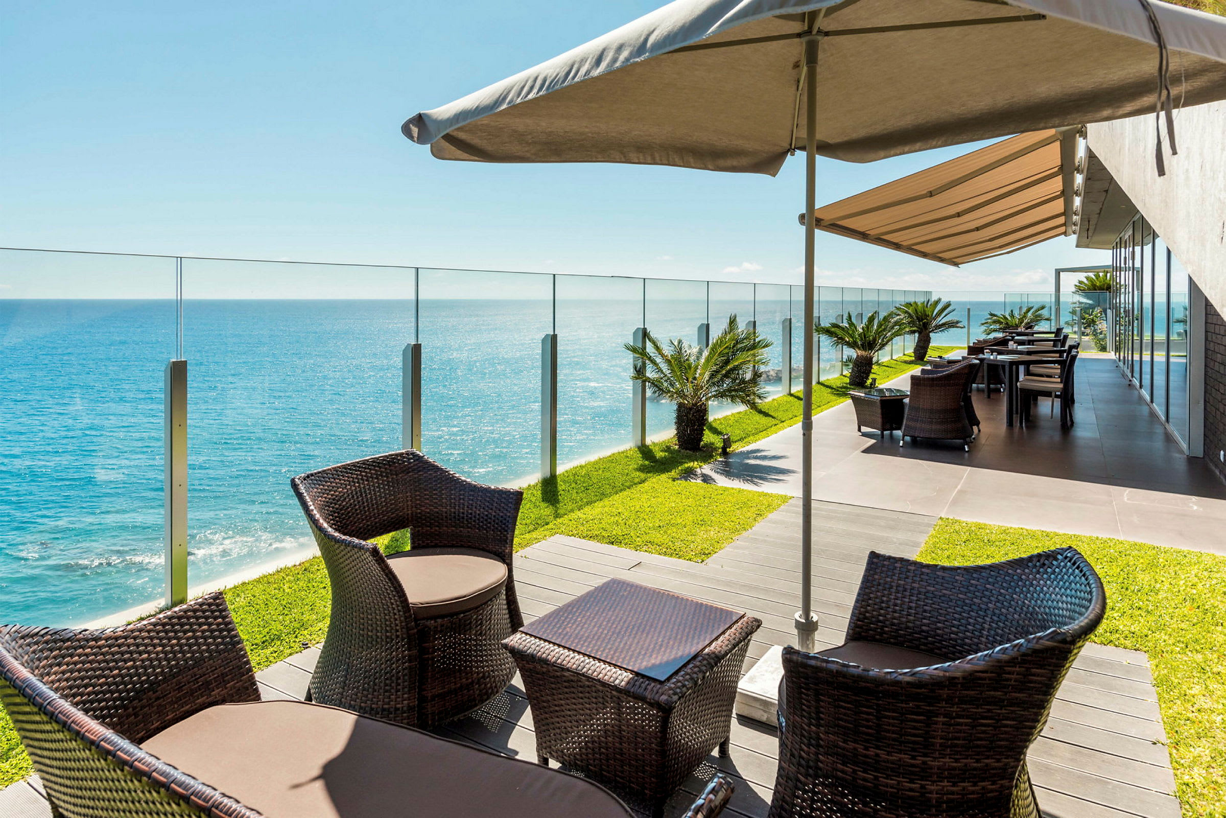 Terrasse med siddepladser, parasol og glasrækværk med udsigt over havet. (AI-genereret tekst)