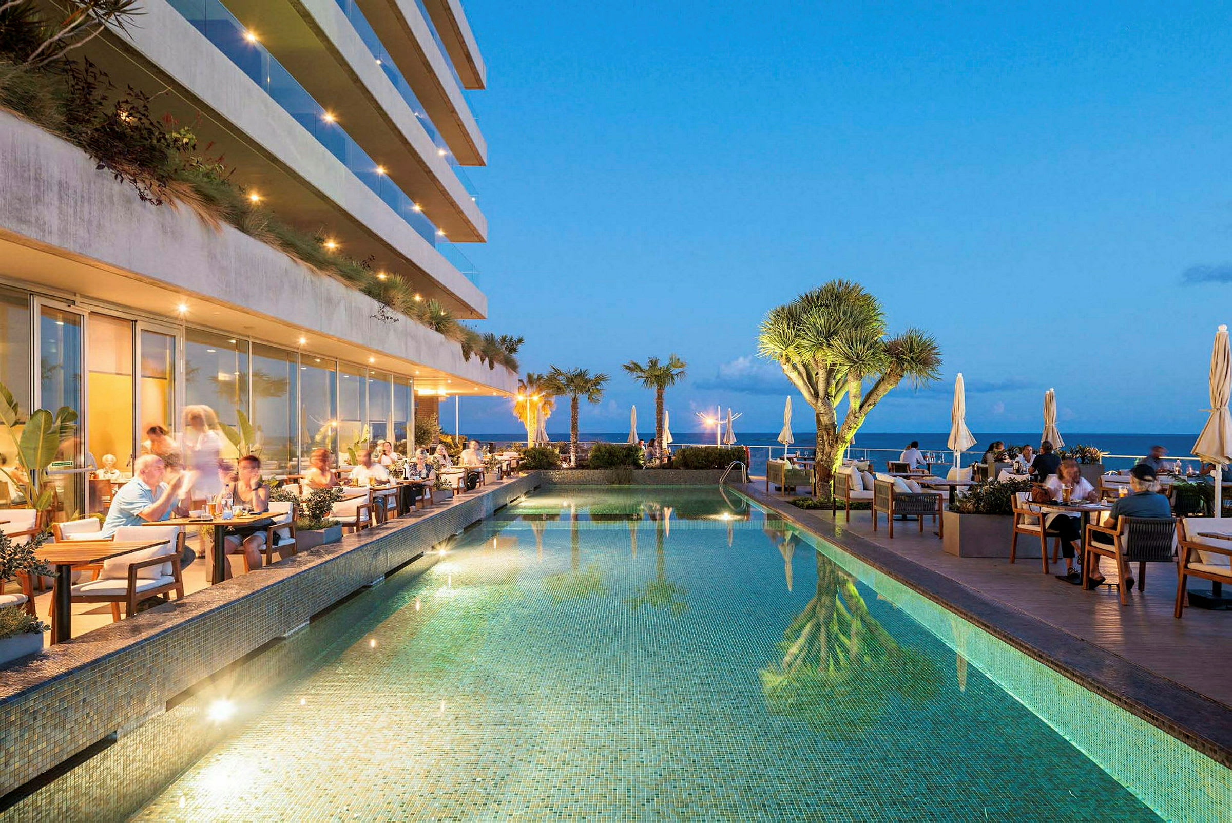 En restaurant med siddepladser ved siden af en pool med udsigt over havet og omkringliggende planter. (AI-genereret tekst)