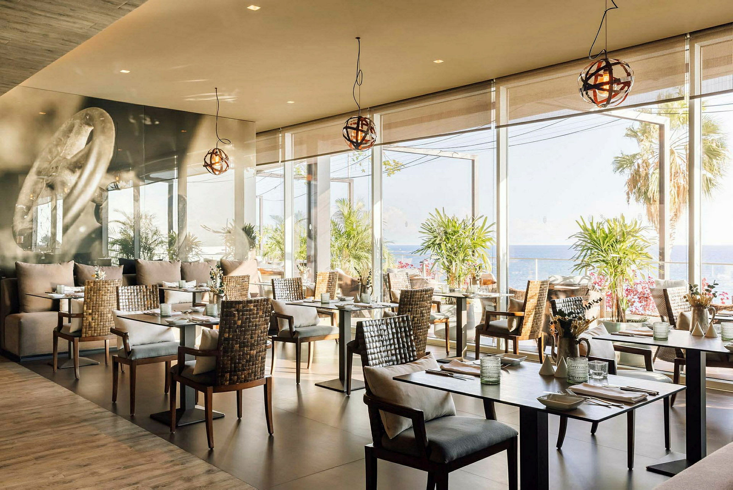 En restaurant med moderne borde og stole, store vinduer med udsigt over havet og palmer. (AI-genereret tekst)
