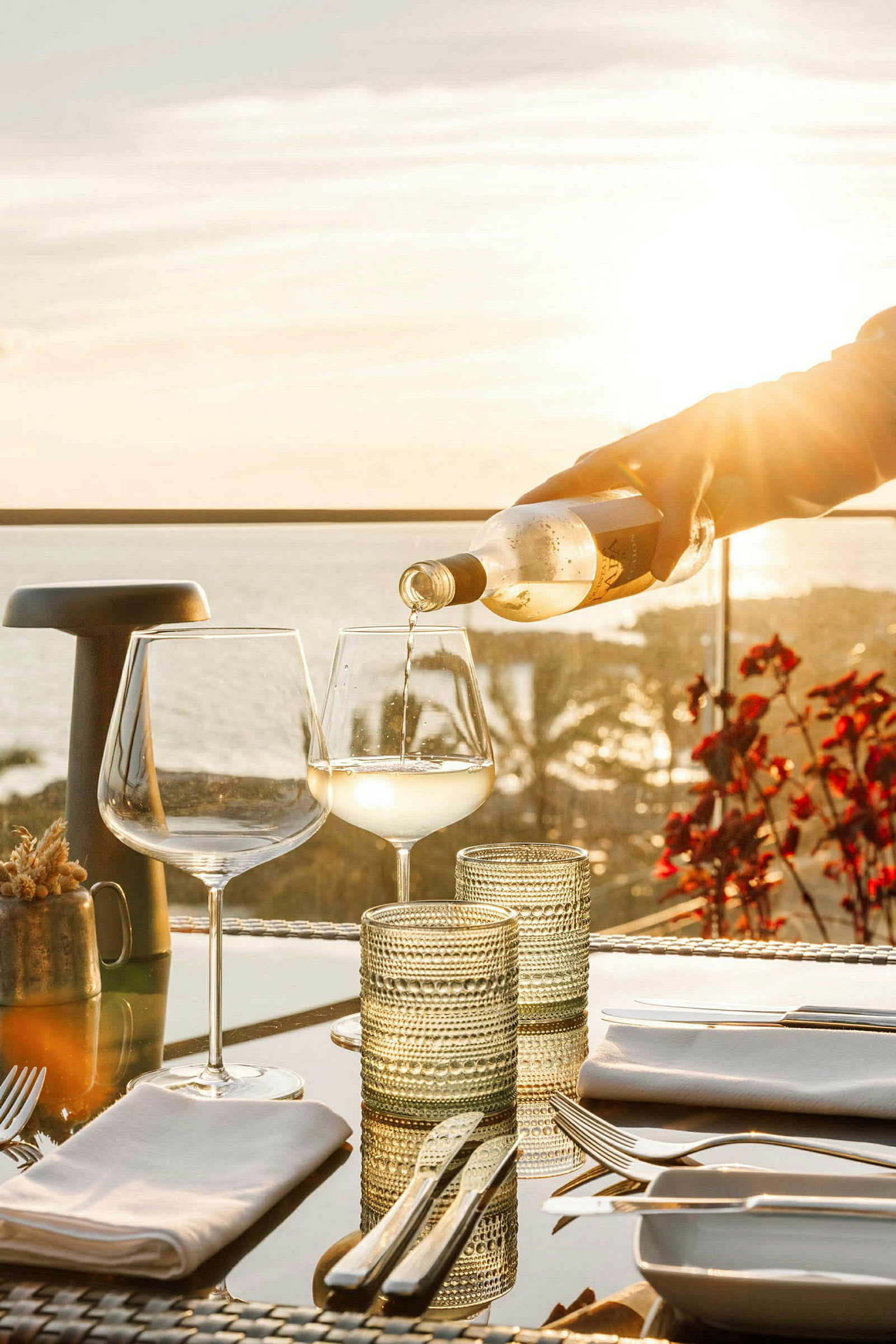 Hvid drik hældes i et vinglas på et bord med redskaber og glas, med solnedgang og hav i baggrunden. (AI-genereret tekst)