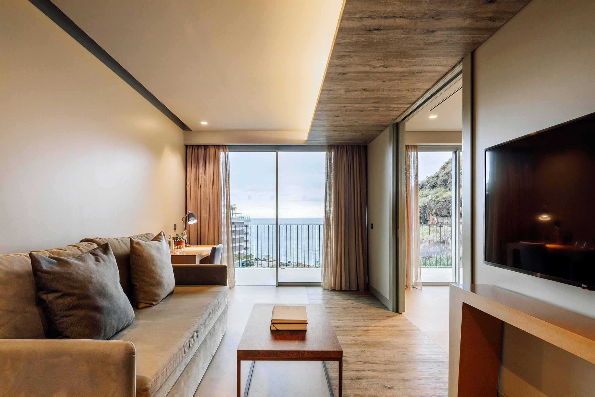 Et hotelværelse med sofa, bord, skrivebord og udsigt over havet gennem store vinduer. (AI-genereret tekst)