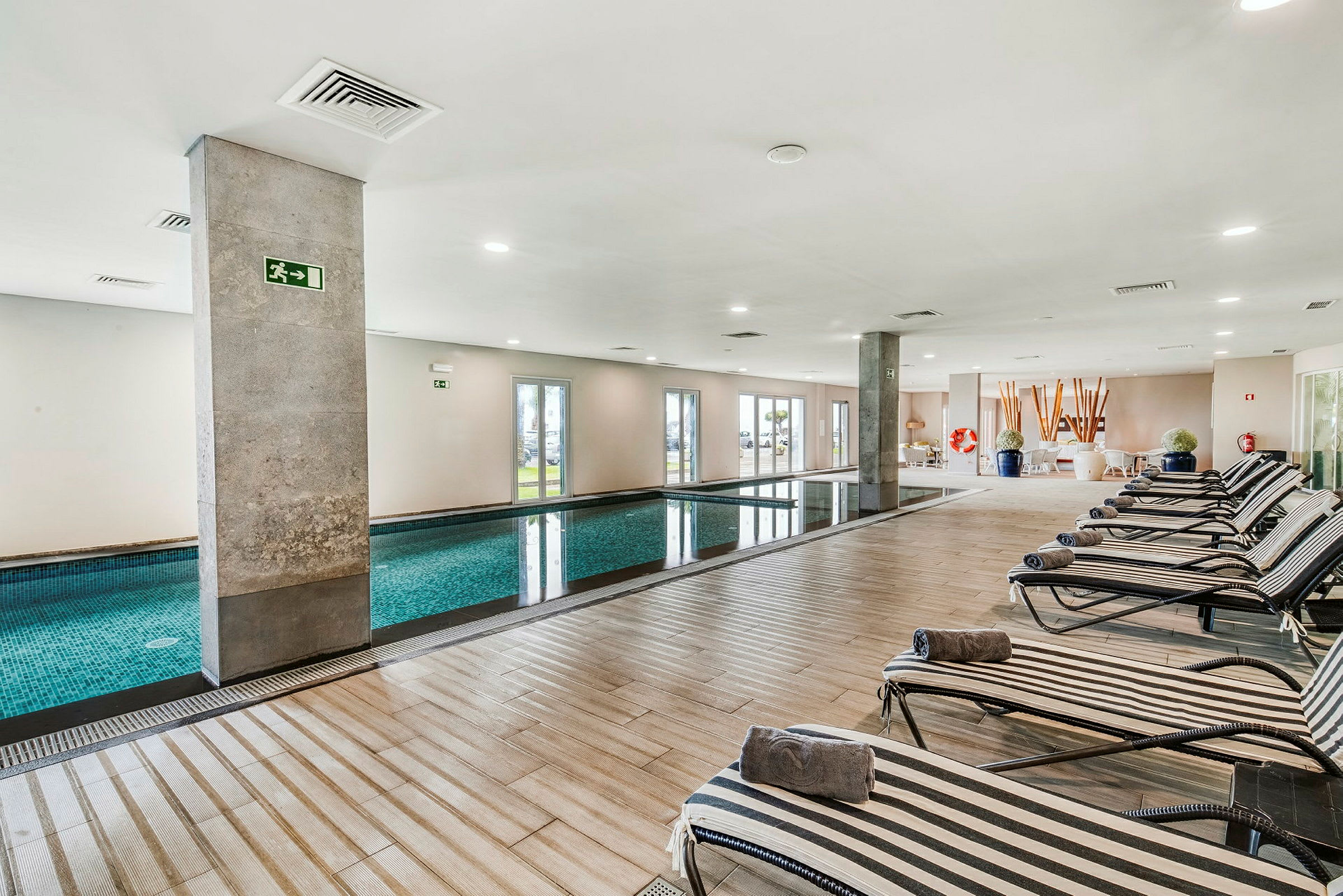Billede af en indendørs pool med liggestole langs den ene side. Rummet er lyst med store vinduer og moderne indretning. (AI-genereret tekst)