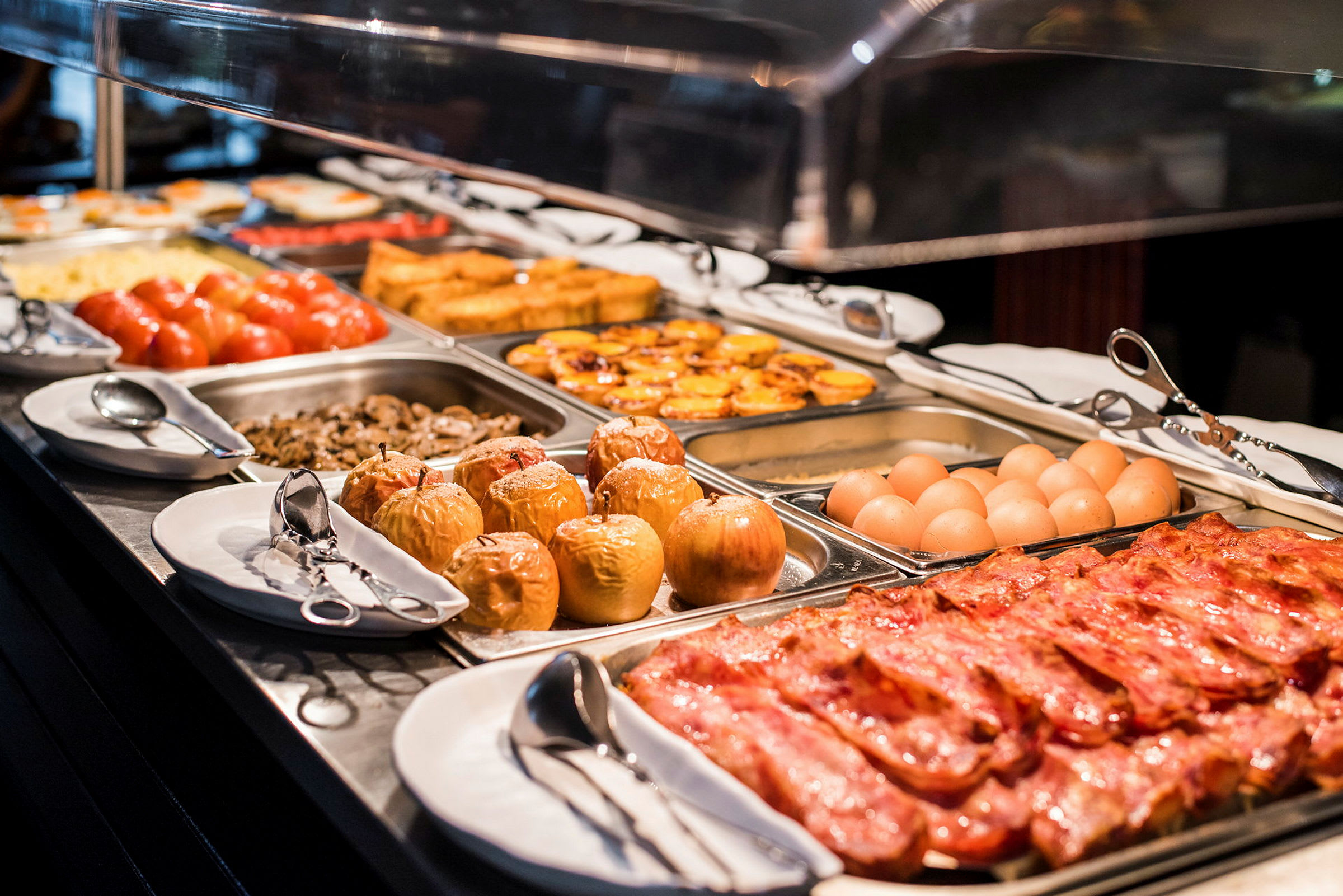 Et buffetbord med forskellige retter, herunder bacon, æg, bagte æbler, tomater og gratiner. (AI-genereret tekst)