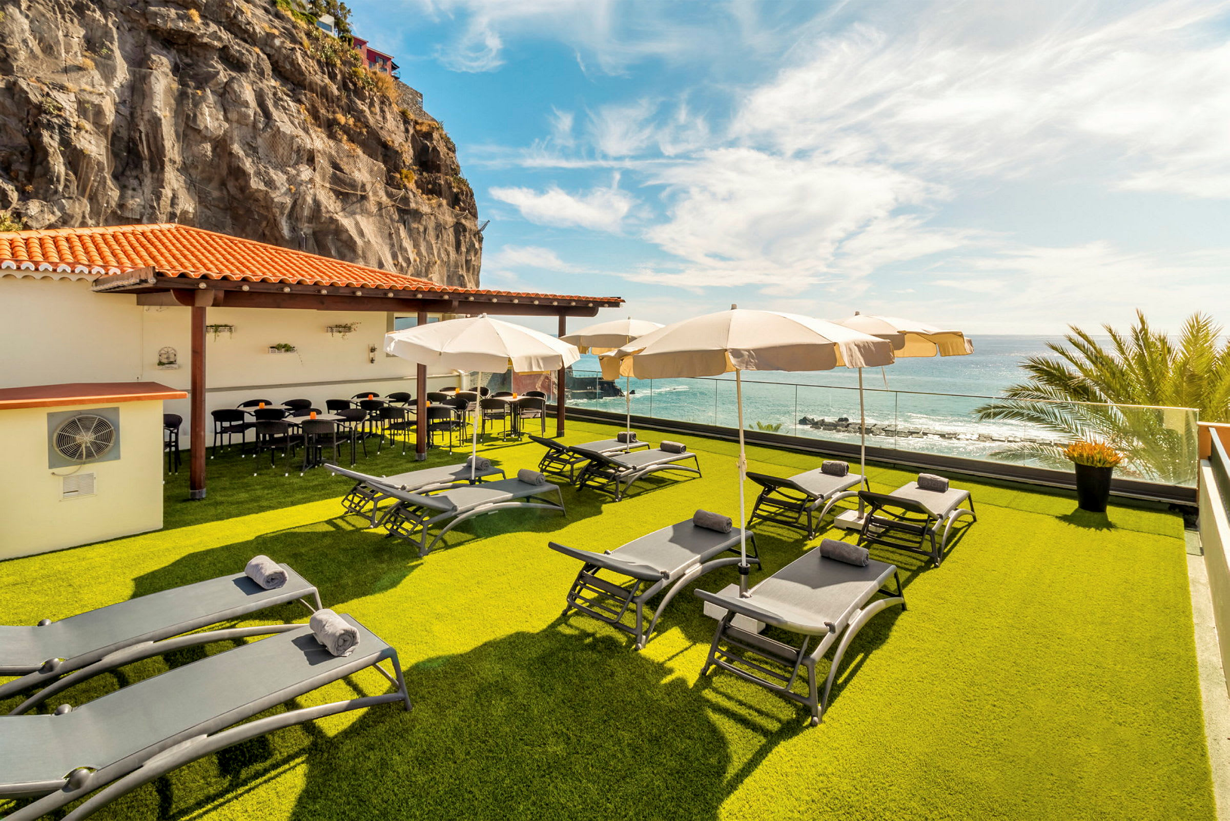 En terrasse med liggestole og parasoller på en græsplæne, ved et lavt hus med et orange tegltag. Udsigt over havet og en klippevæg i baggrunden. (AI-genereret tekst)