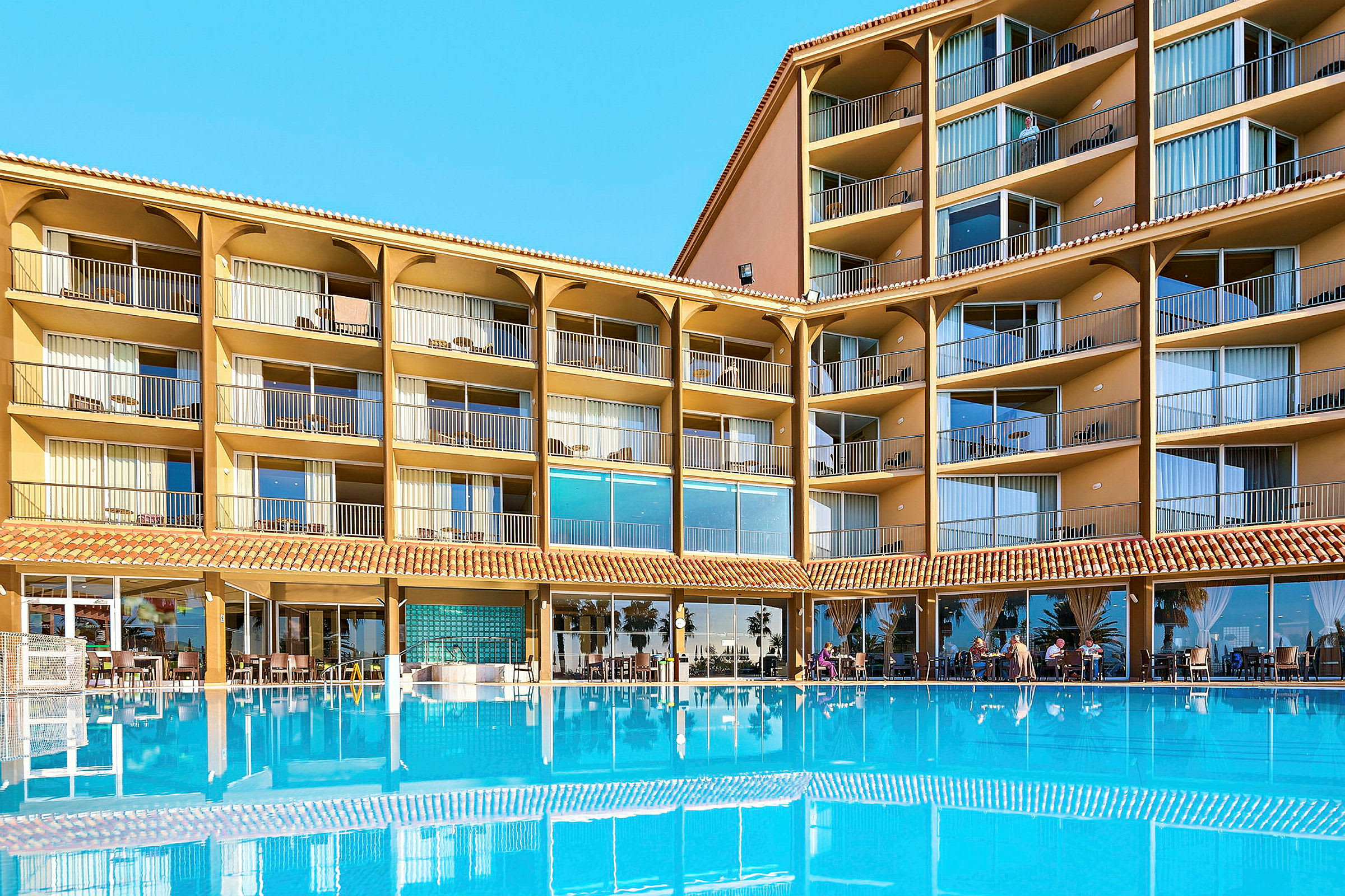 Hotelbygning med flere etager og altaner, foran en stor blå pool. (AI-genereret tekst)