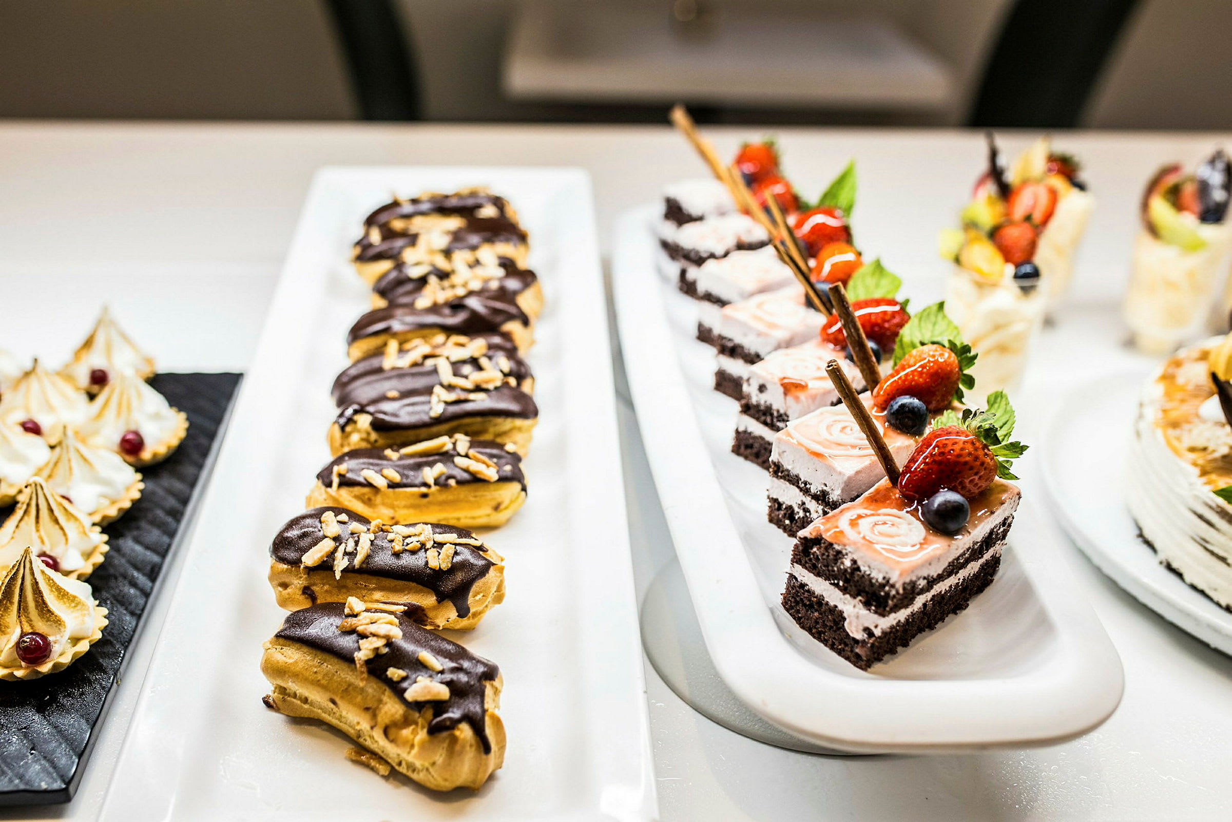Et bord med forskellige desserter, herunder chokoladeovertrukne eclairs og kagebidder med frugtdekoration. (AI-genereret tekst)