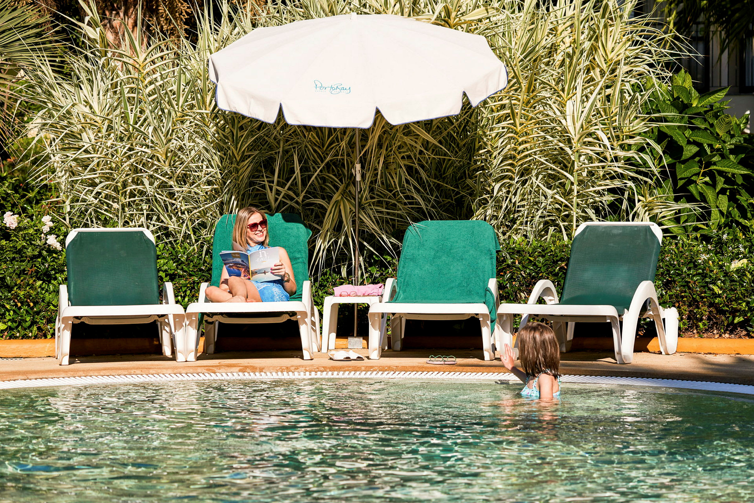 To liggestole ved en pool, en person sidder og læser under en parasol, mens en anden er i poolen. (AI-genereret tekst)