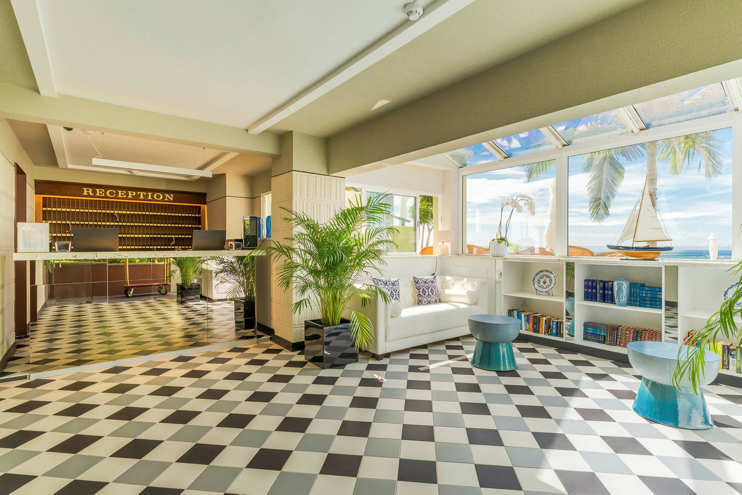 Hotelreception med sort-hvidt gulv, en indbydende sofa og dekorative elementer som en modelsejlskib og bøger i reolen samt udsigt over palmer og havet gennem brede vinduer. (AI-genereret tekst)