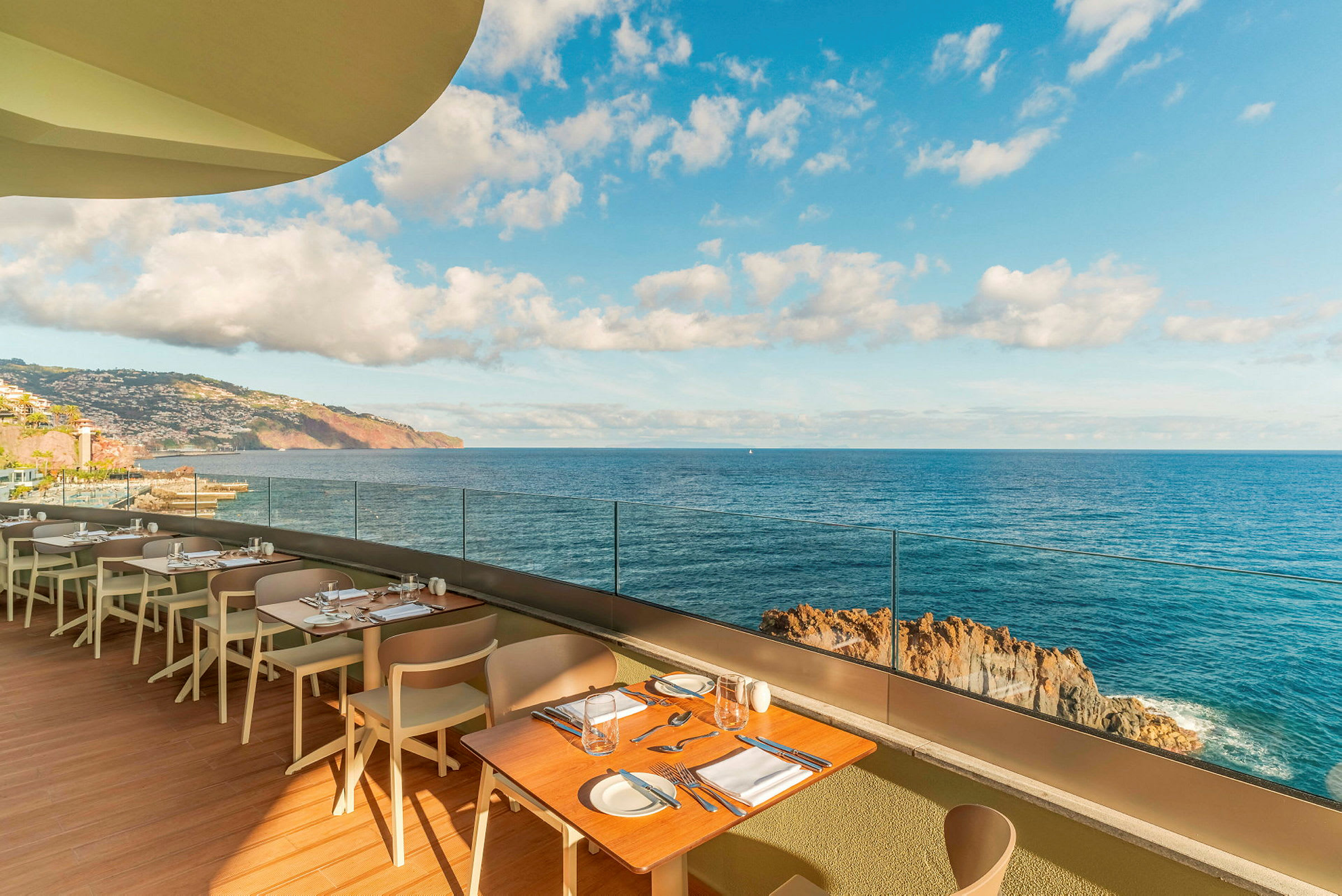Restaurantterrasse med borde og stole, med udsigt over havet og en klippet kyst under en klar himmel. (AI-genereret tekst)