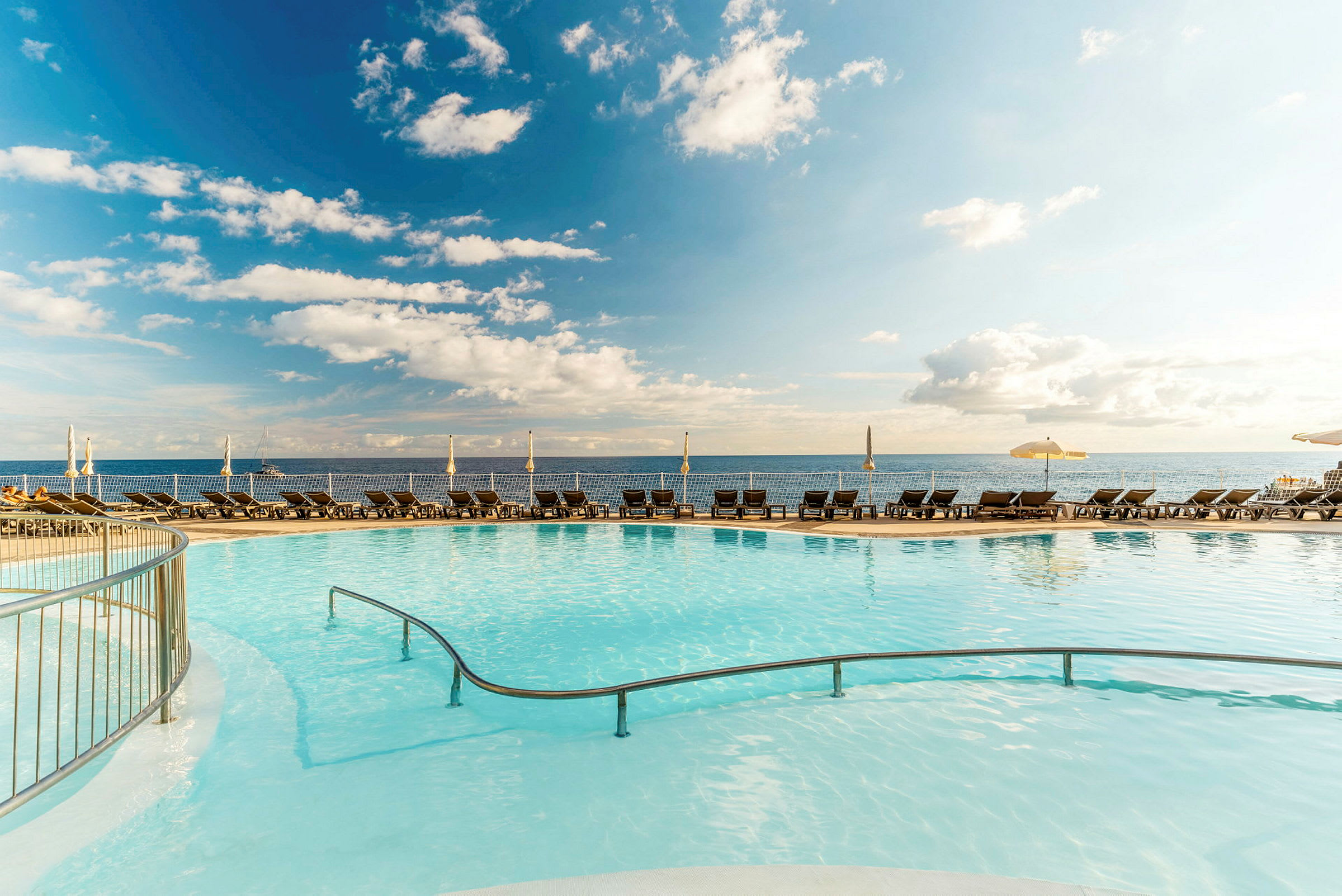 En pool med klart vand, omgivet af liggestole og parasoller, med havet og himlen i baggrunden. (AI-genereret tekst)