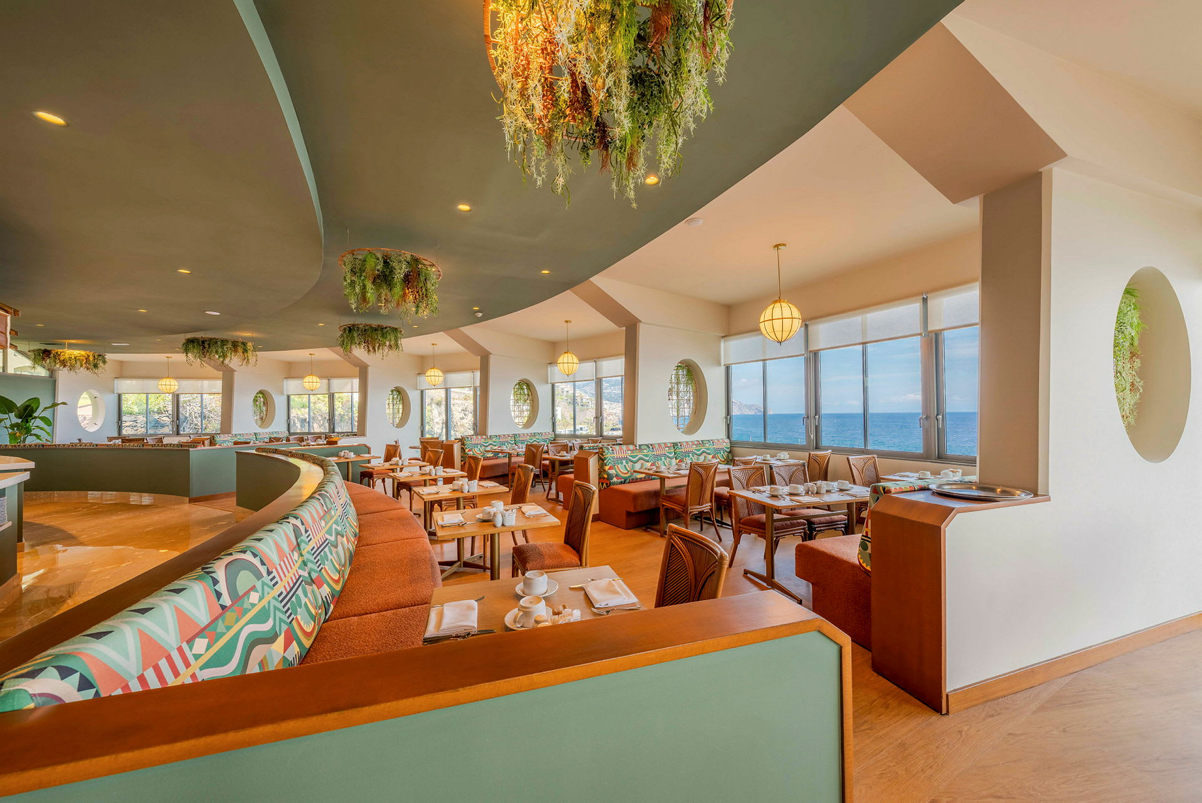 Restaurant med store vinduer og udsigt over havet, indrettet med farverige sofaer og grønne dekorationer. (AI-genereret tekst)