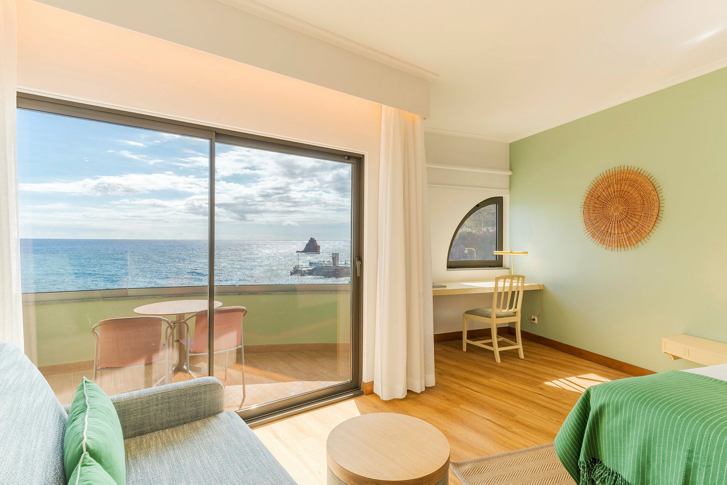 Et hotelværelse med udsigt over havet gennem store glasdøre. På verandaen er der en siddegruppe, og interiøret er lyst og moderne. (AI-genereret tekst)