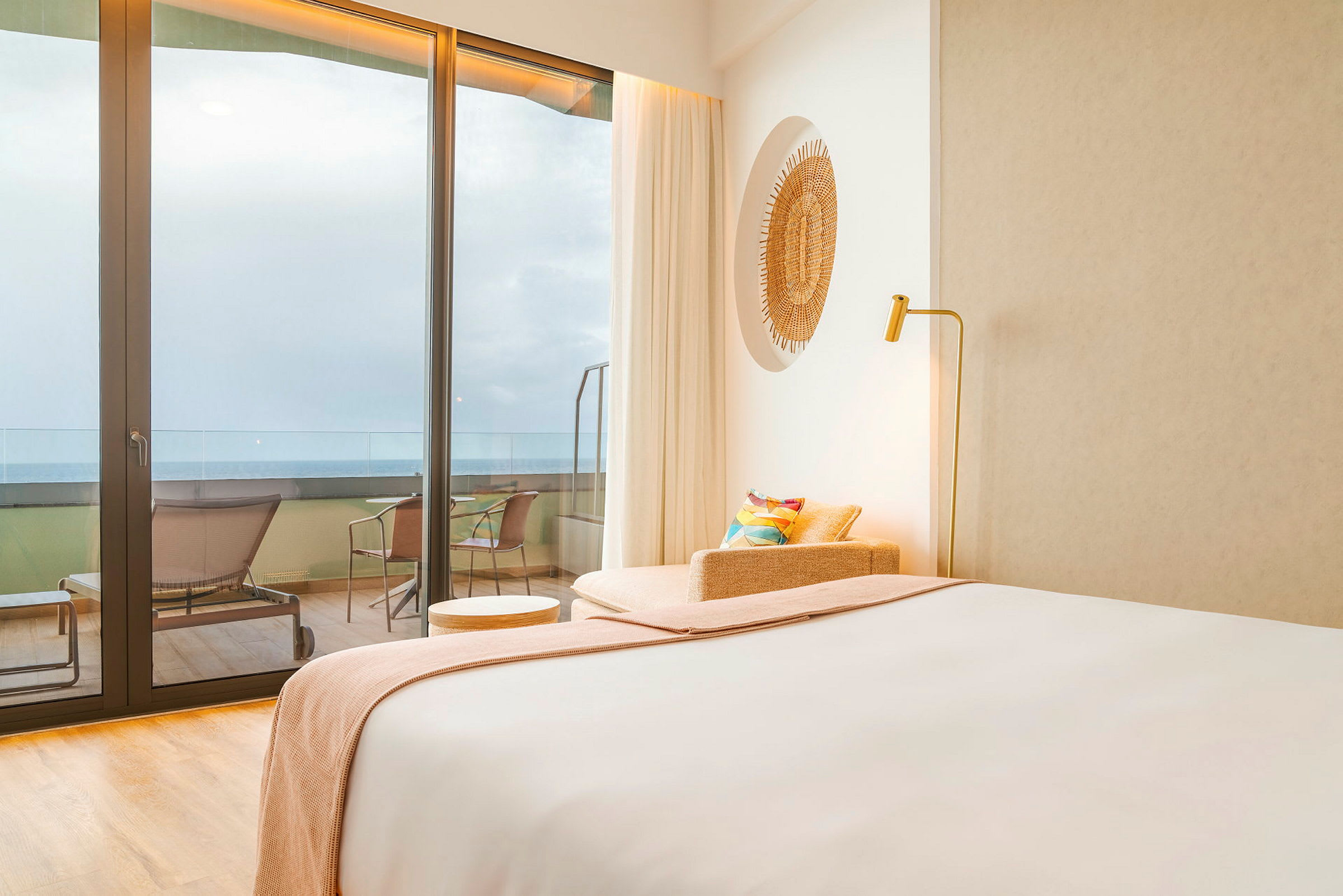 Et hotelværelse med moderne indretning. Der er en seng, en lænestol og et lille bord tæt på en glasflade med udsigt over en altan og havet. (AI-genereret tekst)