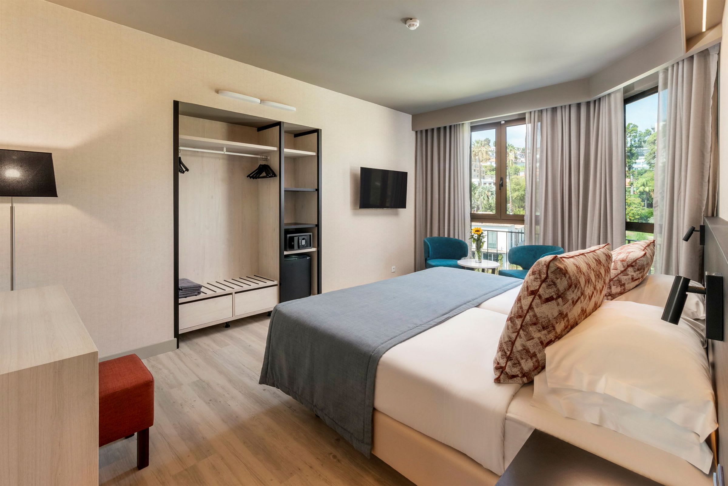 Et hotelværelse med dobbeltseng, to blå stole ved et bord foran vinduer samt garderobe med åbne hylder. (AI-genereret tekst)