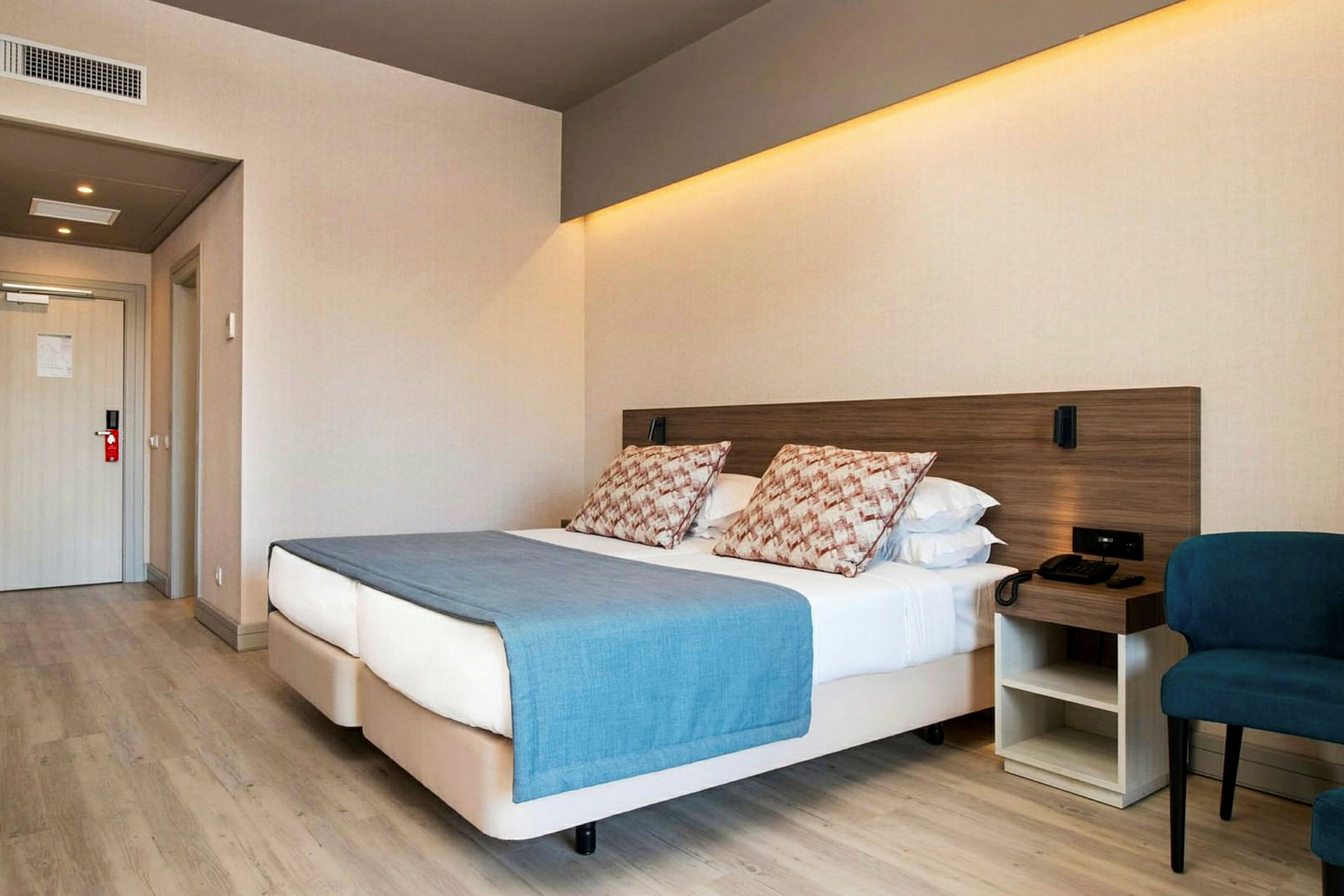 Hotelværelse med en dobbeltseng, blå sengelagen og puder, ved siden af en stol og et natbord. (AI-genereret tekst)