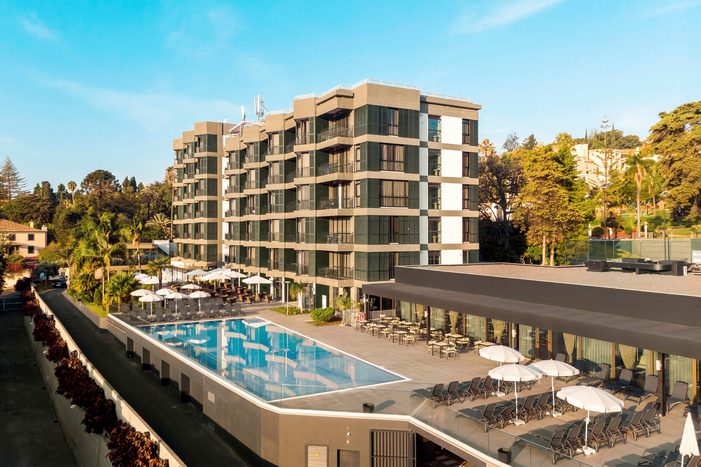Billede af et hotel med udendørs pool og liggestole, omgivet af træer og vegetation. (AI-genereret tekst)