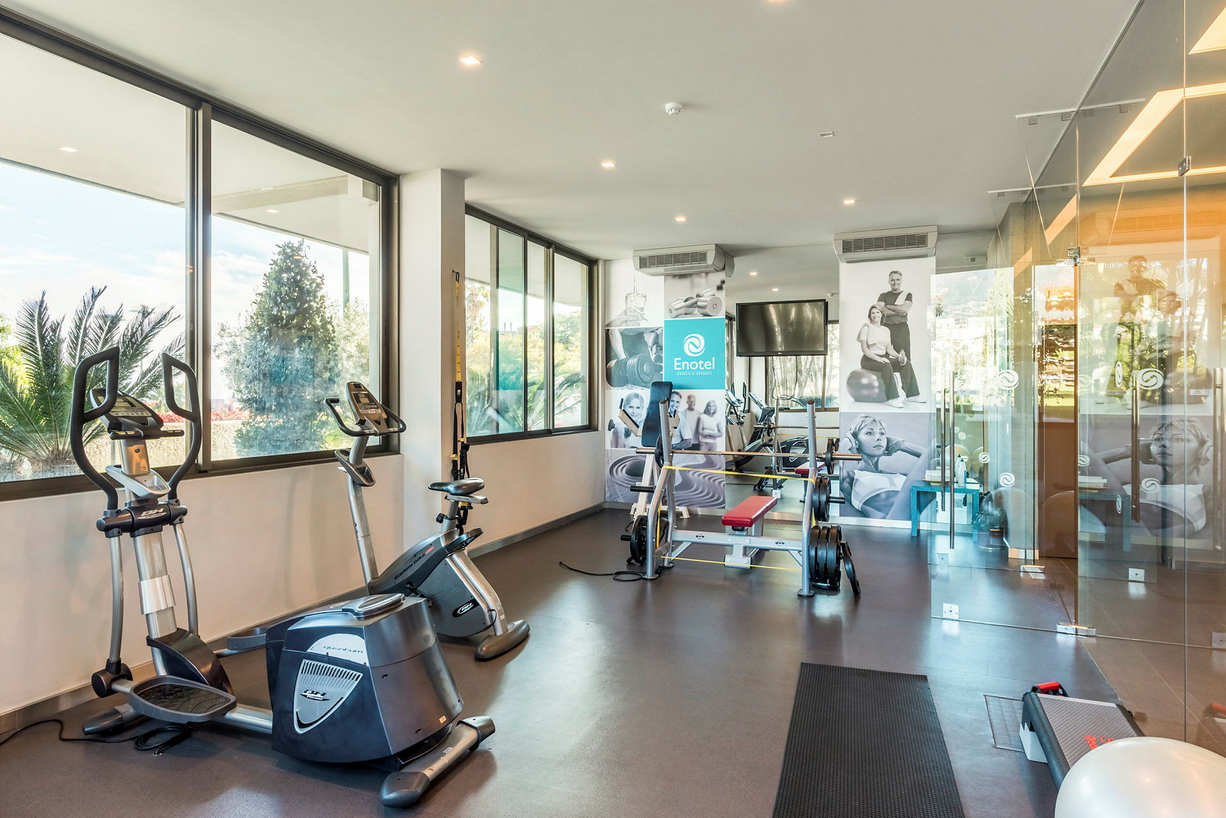 Et fitnesscenter med træningsudstyr inklusive motionscykler og styrketræningsmaskiner, med store vinduer der lukker naturligt lys ind. (AI-genereret tekst)