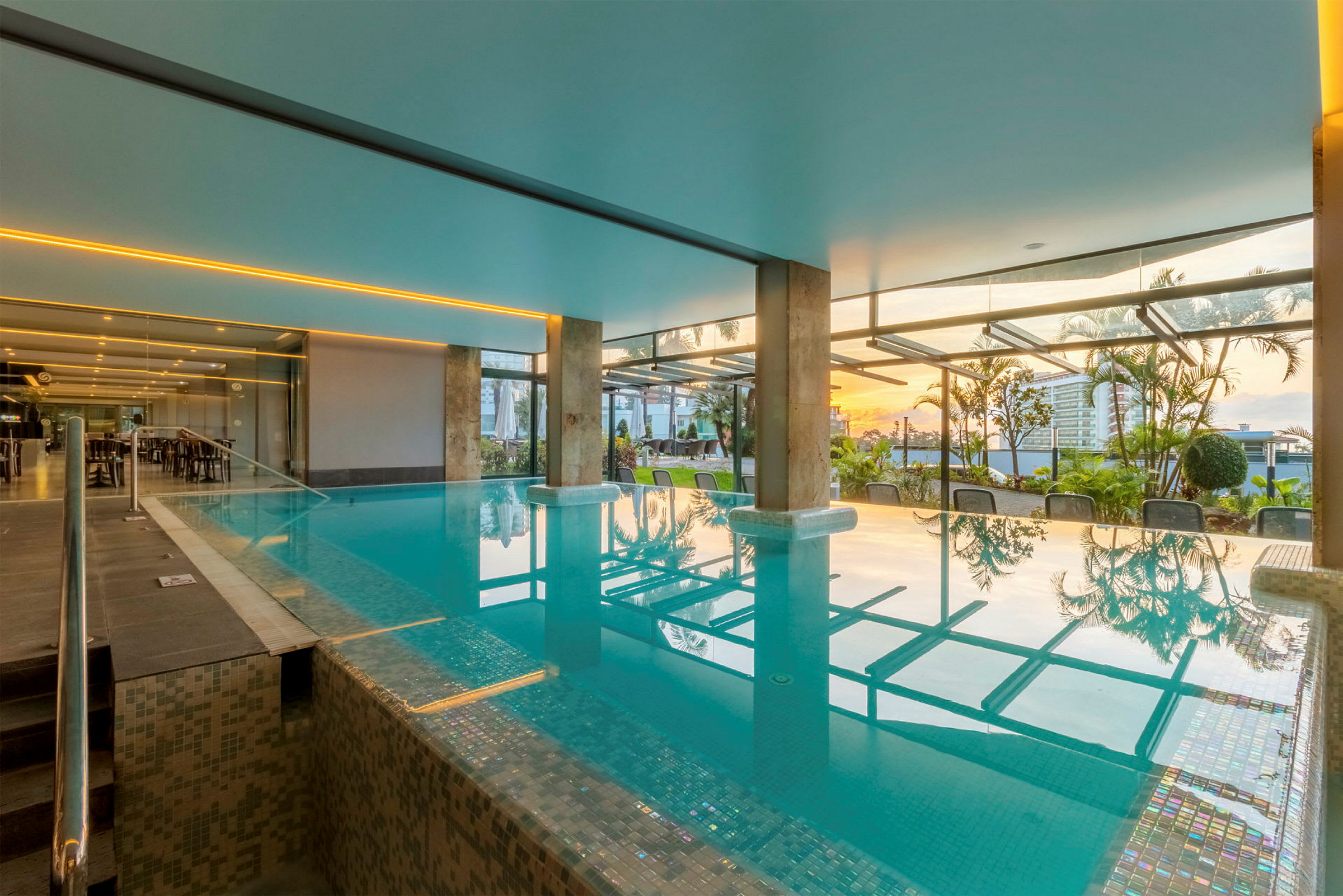 Moderne indendørs swimmingpool med store glasvinduer og udsigt til have og solnedgang. (AI-genereret tekst)