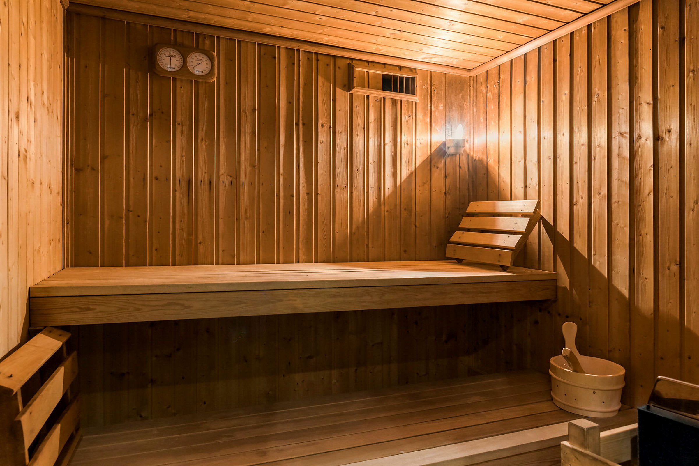 Indretning af en sauna med træpaneler, siddebænk, øse og saunaventilator. (AI-genereret tekst)