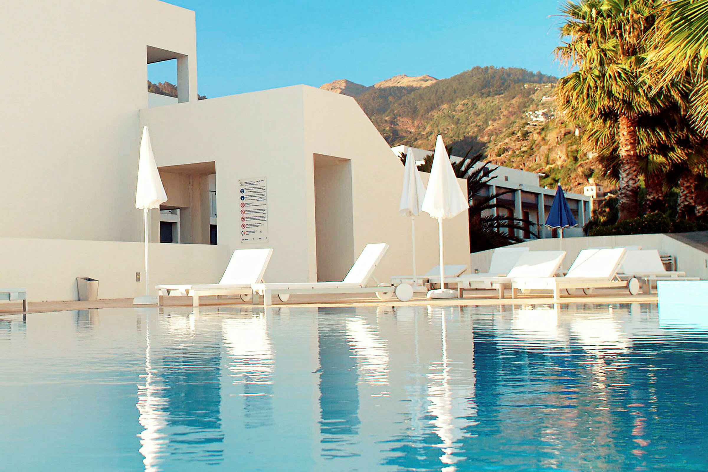 Udendørs swimmingpool med liggestole og parasoller ved siden af en hvid bygning. (AI-genereret tekst)