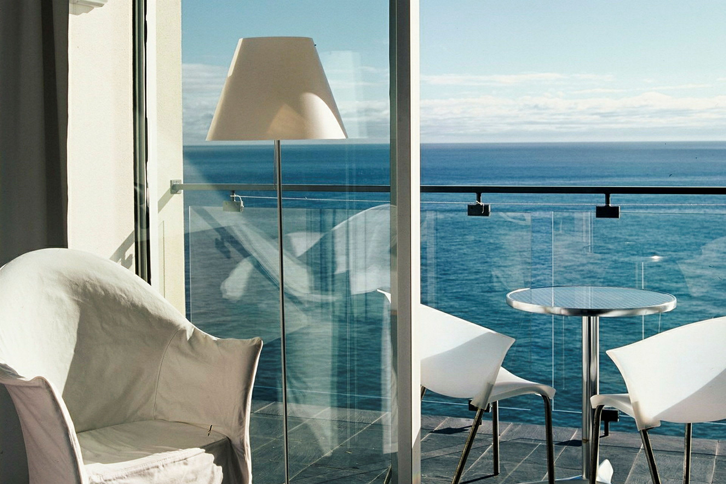 Udsigt fra et værelse mod havet med en balkon med glasrækværk, bord og stole. (AI-genereret tekst)