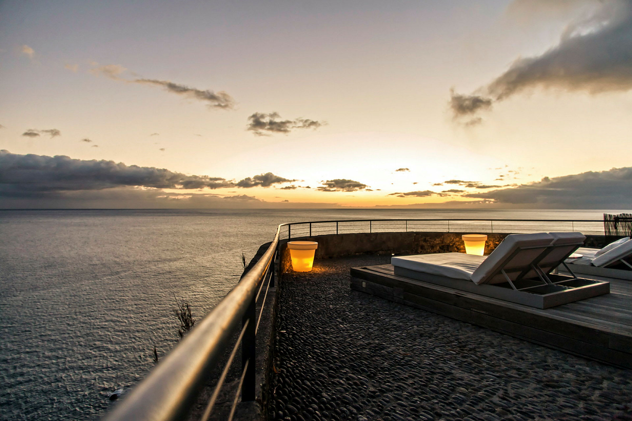 Terrasse ved havet med liggestole og en lampe i solnedgang. (AI-genereret tekst)