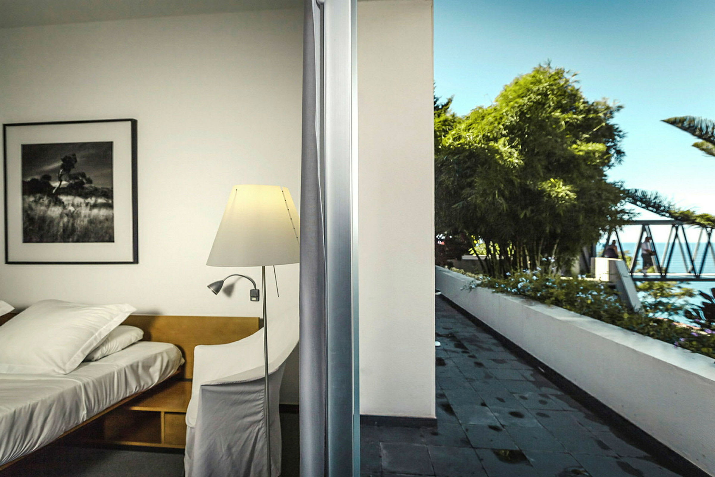 Et værelse med en seng og en læselampe ved siden af en balkon med udsigt til en pool og træer. (AI-genereret tekst)