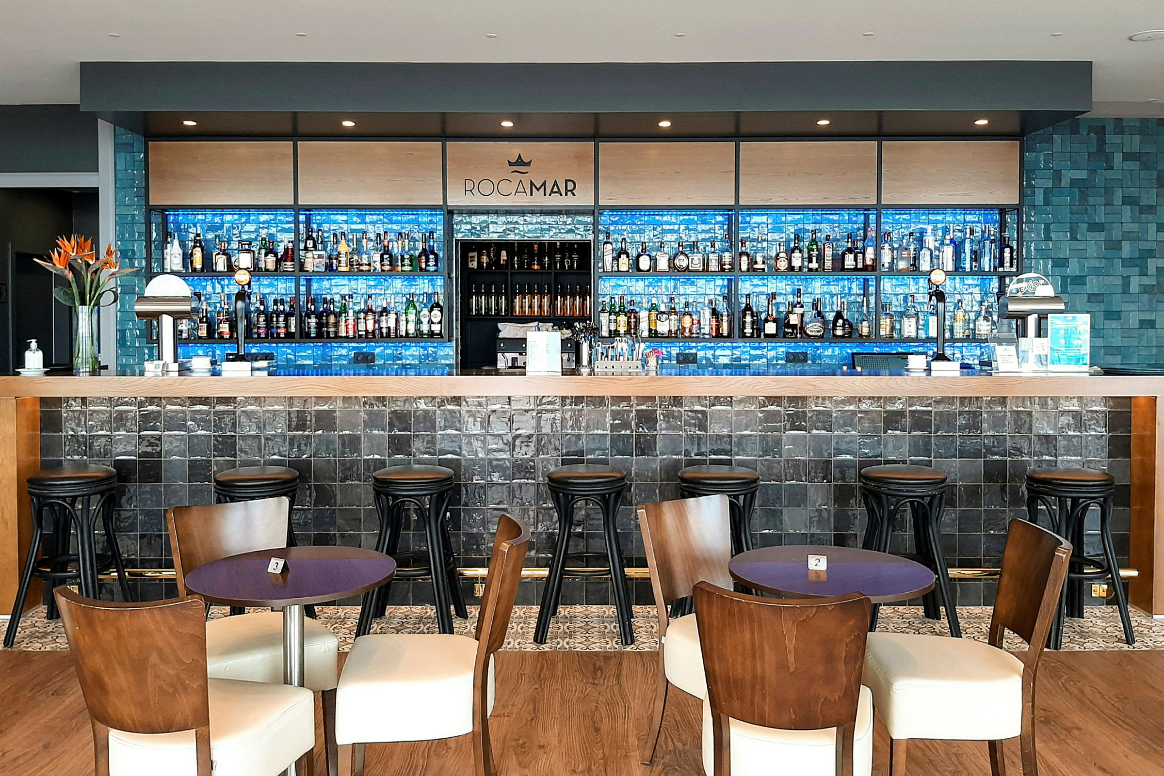 En hotelbar med træborde og stole foran en lang bardisk. (AI-genereret tekst)
