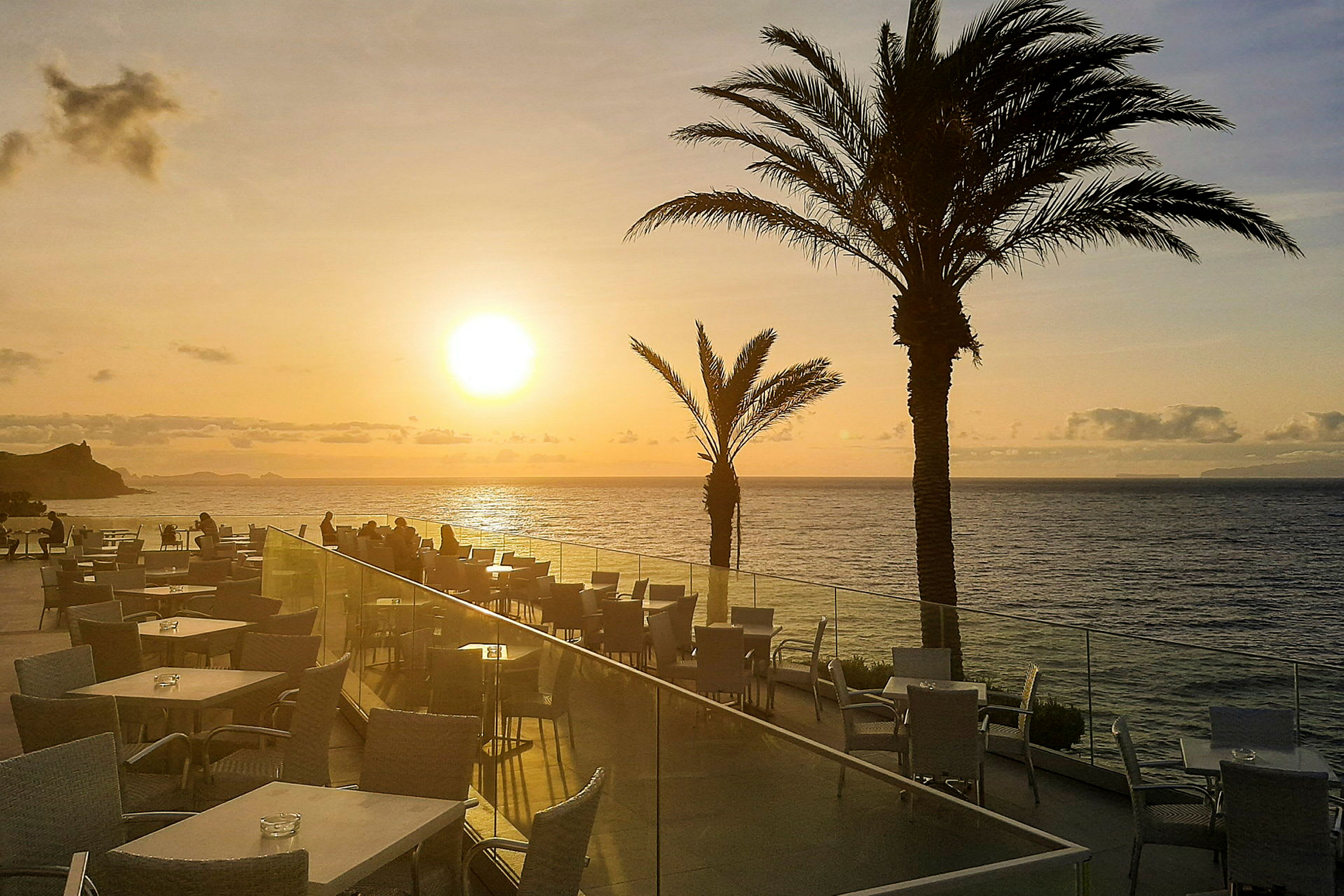 Terrasse med borde og stole, i baggrunden ses havet og en solnedgang. (AI-genereret tekst)
