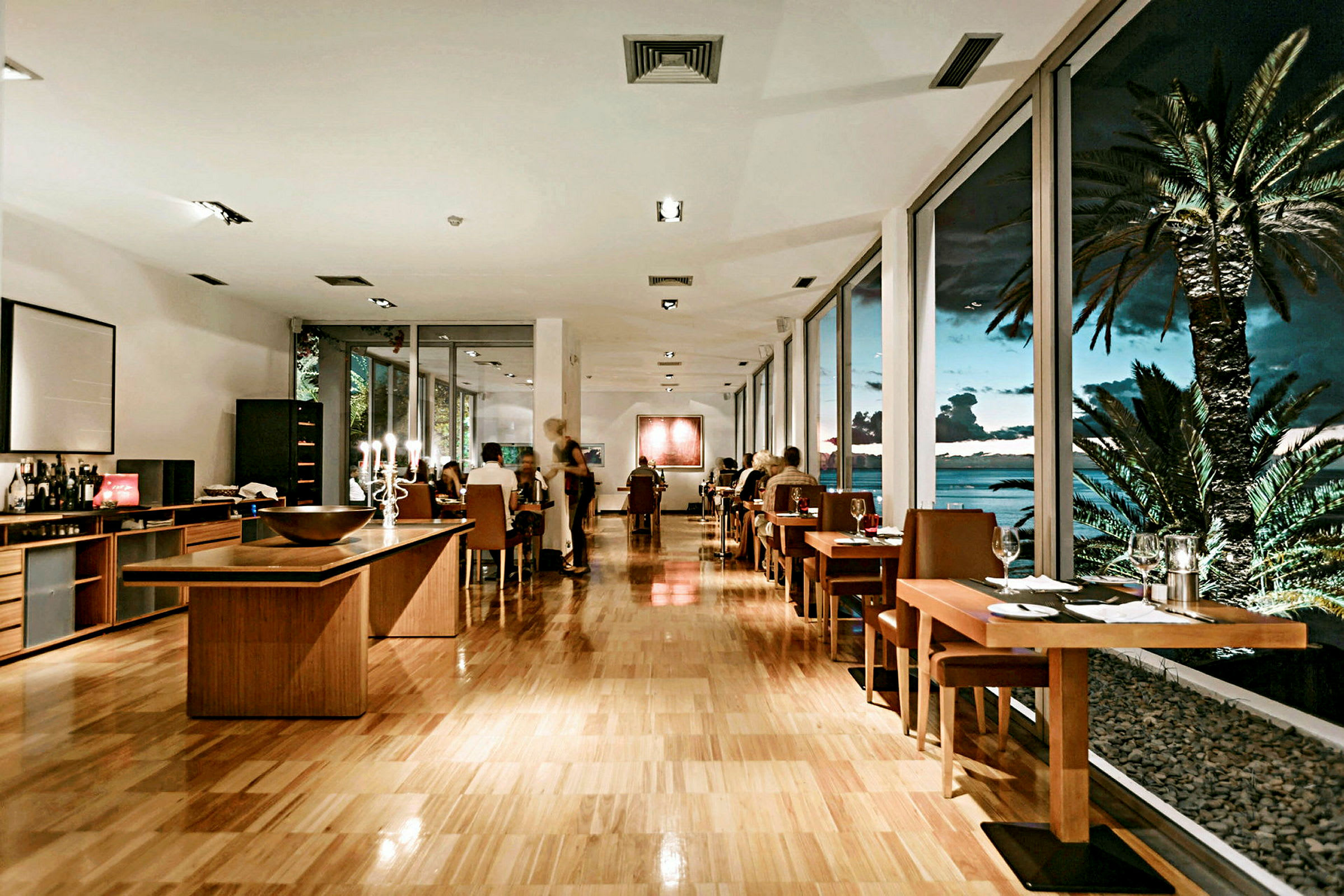 Et elegant indrettet restaurantområde med borde, stole og store vinduer med udsigt over en aftenhimmel med palmer og hav. (AI-genereret tekst)