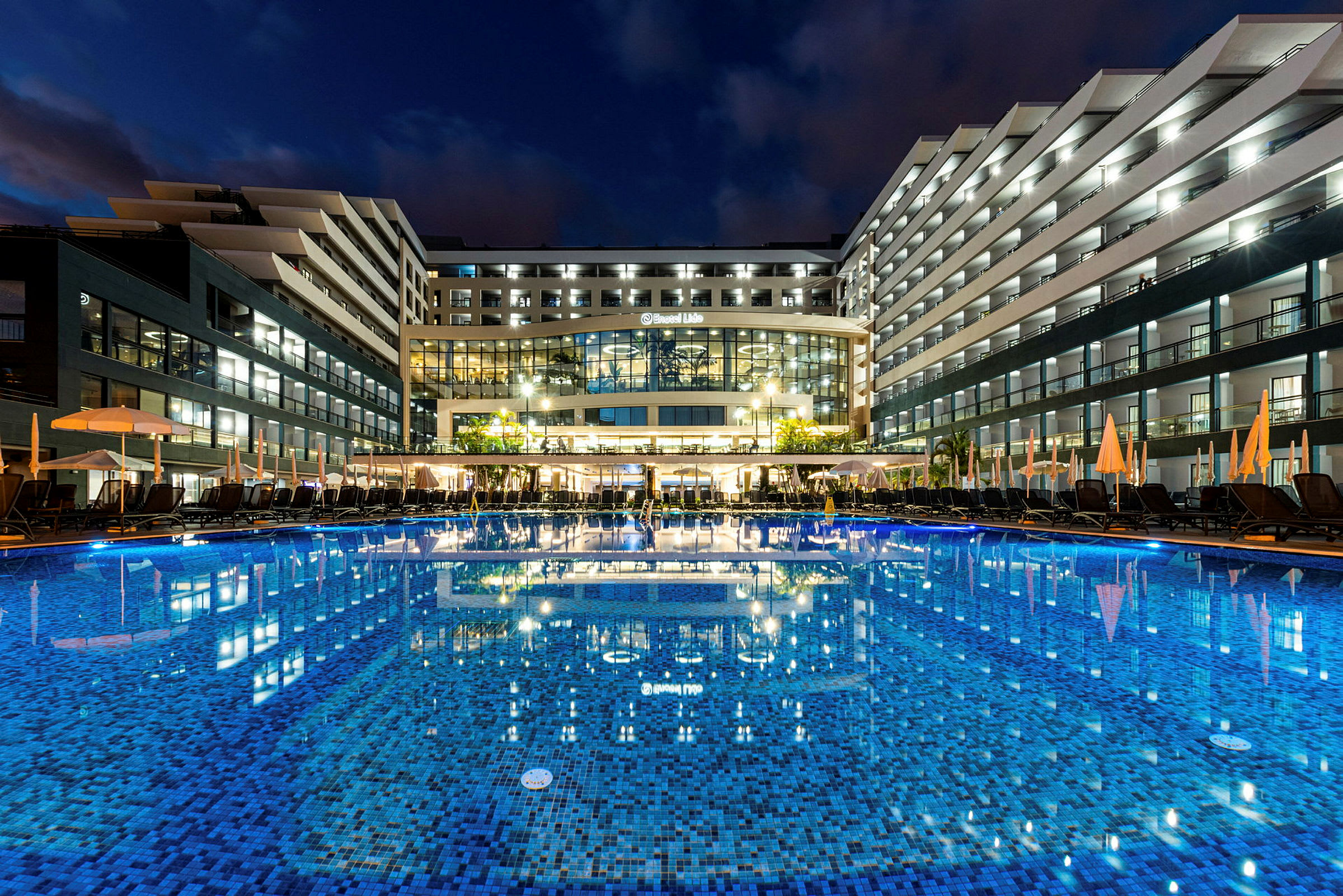 Hotel med oplyste altaner og en stor pool foran, aftenstemning. (AI-genereret tekst)