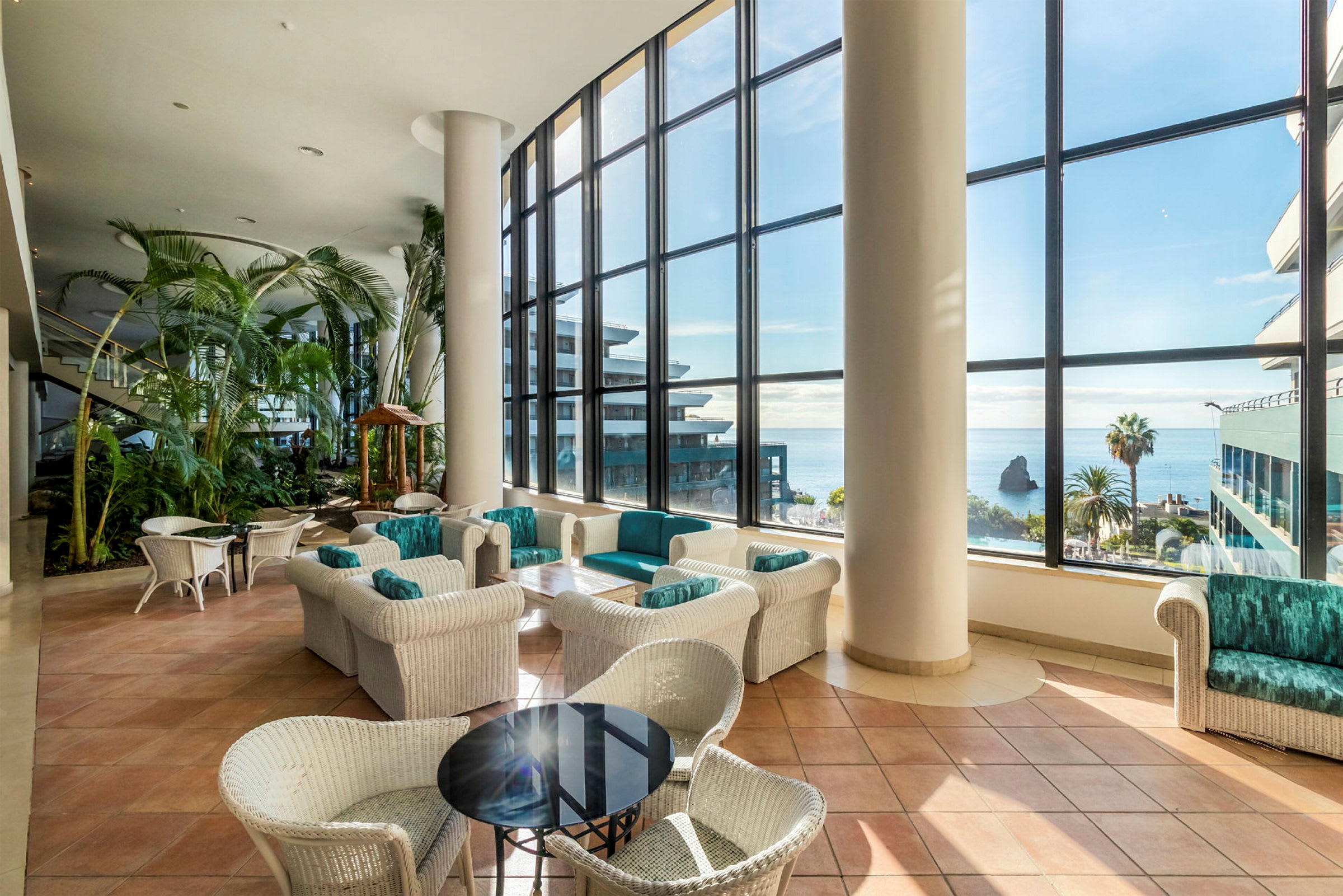 Hotellets lobby med store vinduer ud mod havet. Borde og stole i flettede materialer samt planter. (AI-genereret tekst)