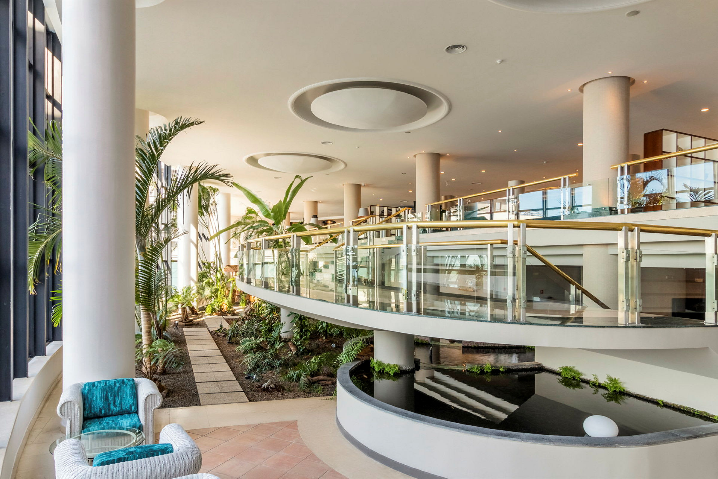 En moderne hotellhal med planter, glasgelændere og vandanlæg. (AI-genereret tekst)