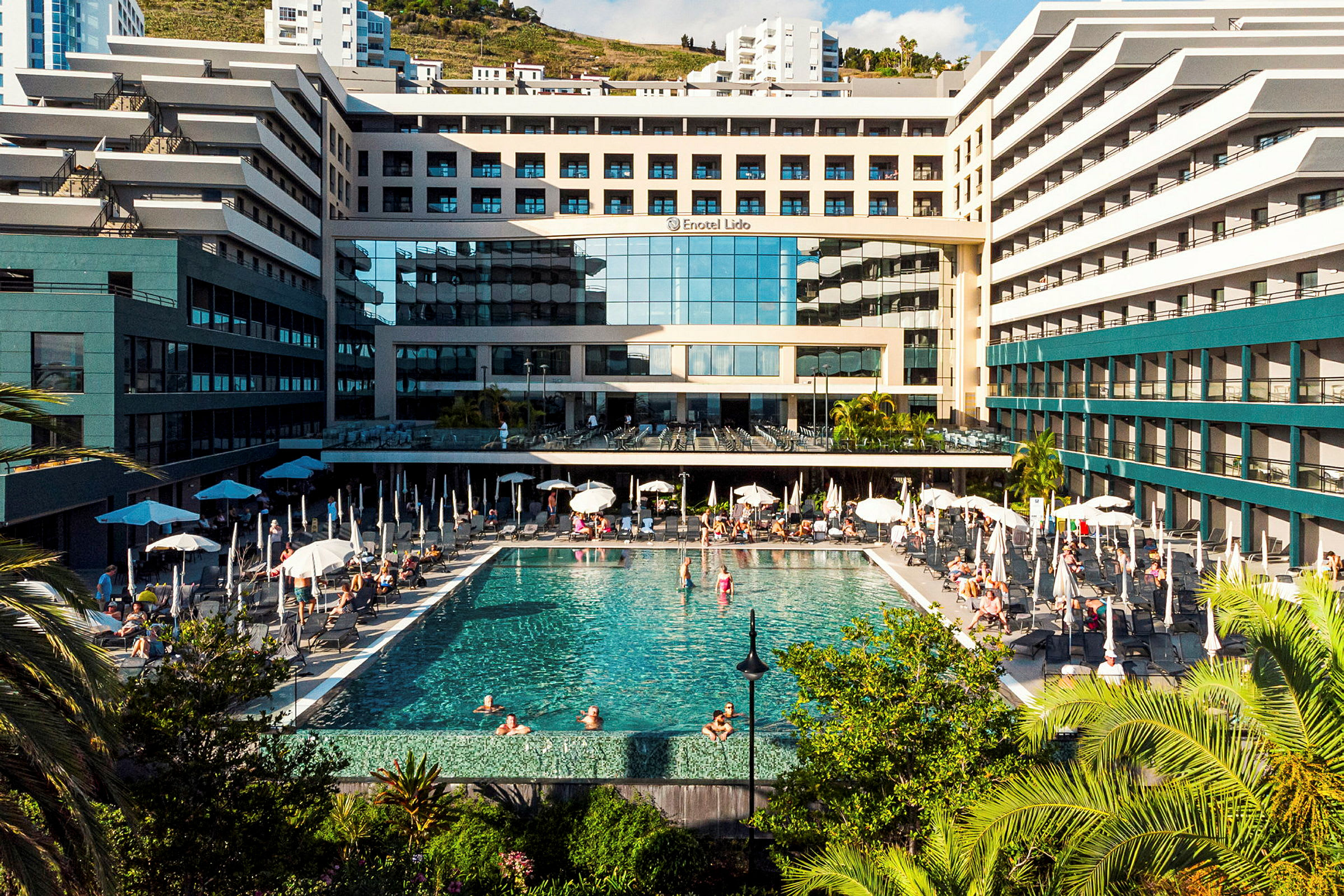 Hotelbygning med en stor udendørs swimmingpool og liggestole, omgivet af vegetation. (AI-genereret tekst)