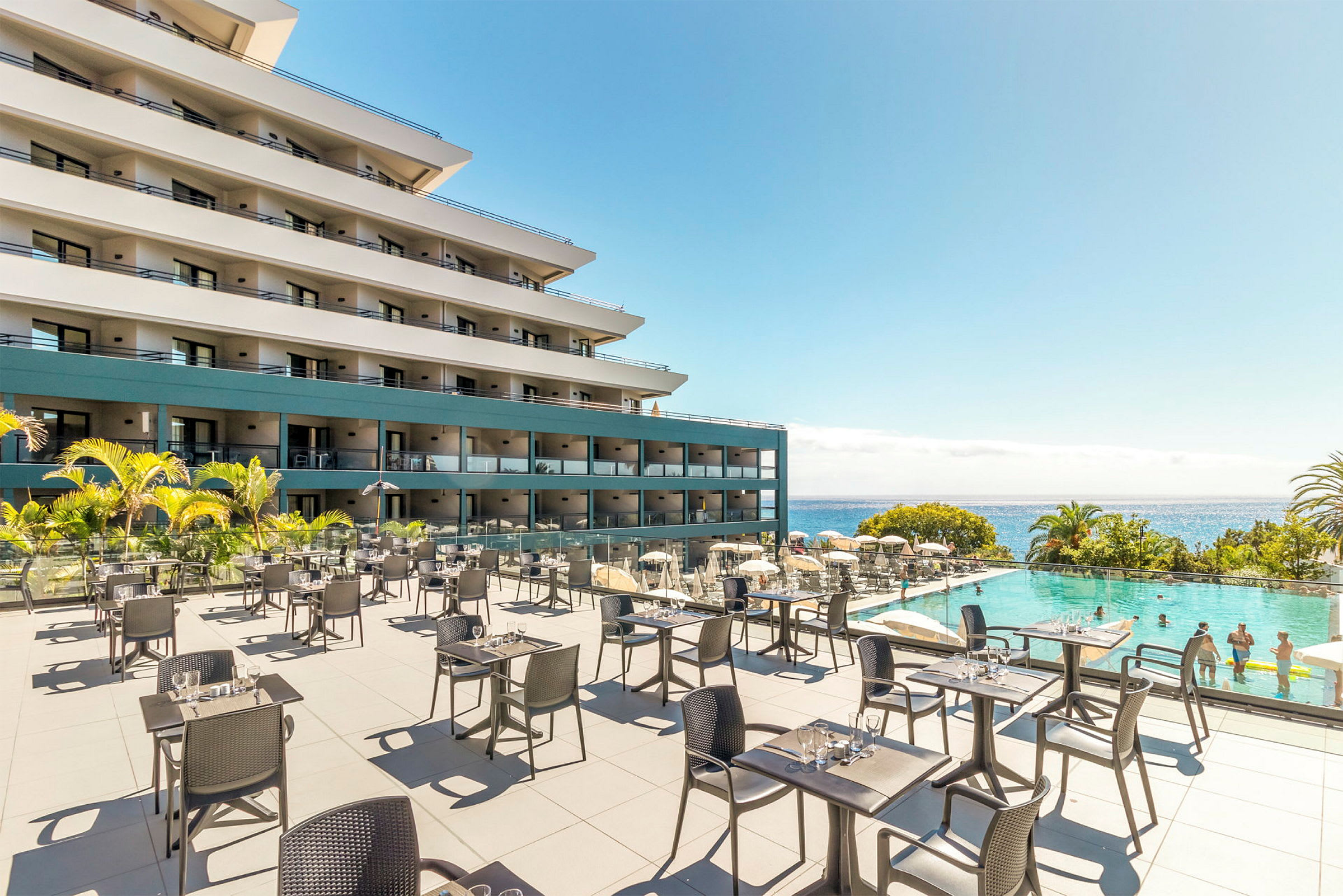 Udendørs serveringsområde med borde og stole foran et hotel og en pool med badende gæster, med havet i baggrunden. (AI-genereret tekst)