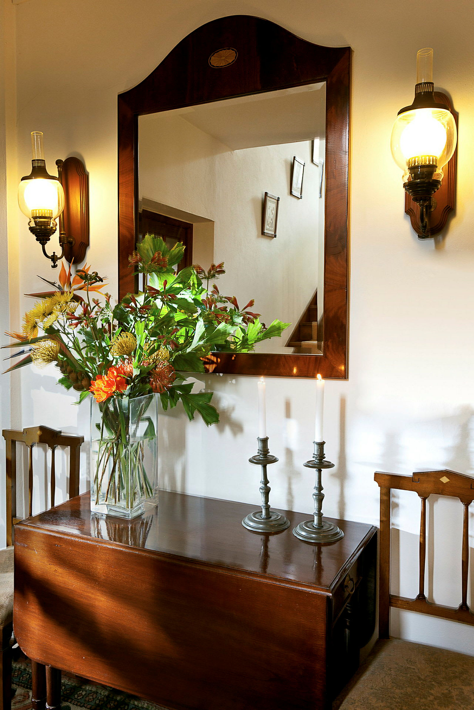 Et spejl med træramme, to væglamper og et bord med en vase med blomster samt to lysestager med tændte lys. (AI-genereret tekst)