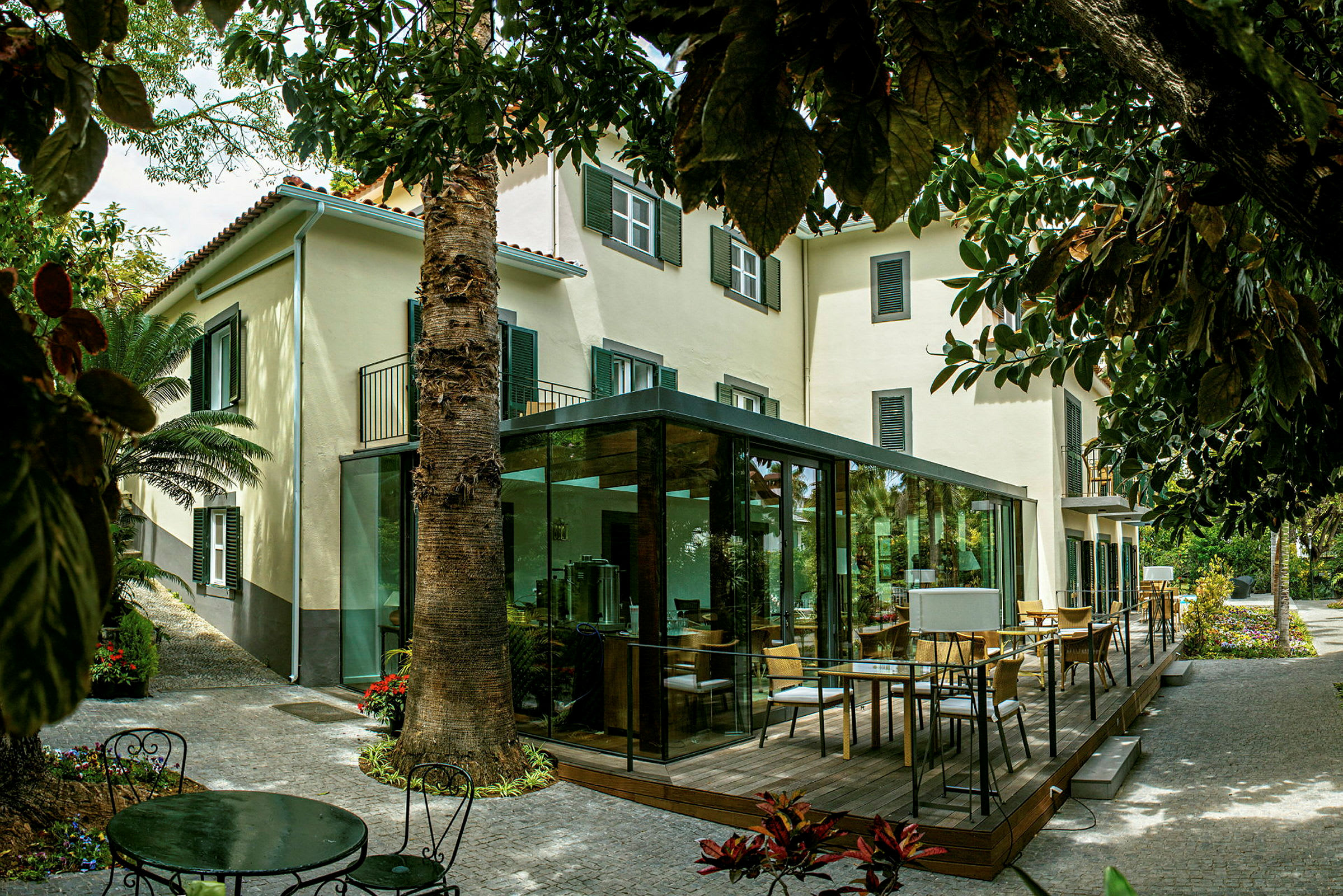 En bygning med beige facade og grønne skodder, omgivet af grønt og med en terrasse med glasvægge. (AI-genereret tekst)