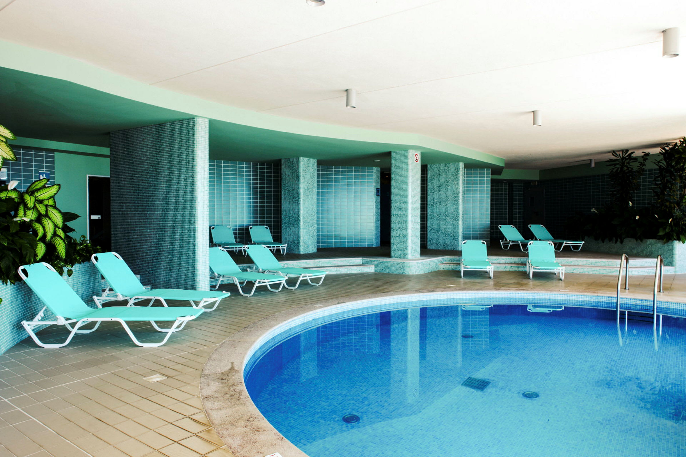 En indendørs pool med blåt vand, omgivet af liggestole og grønne planter. (AI-genereret tekst)