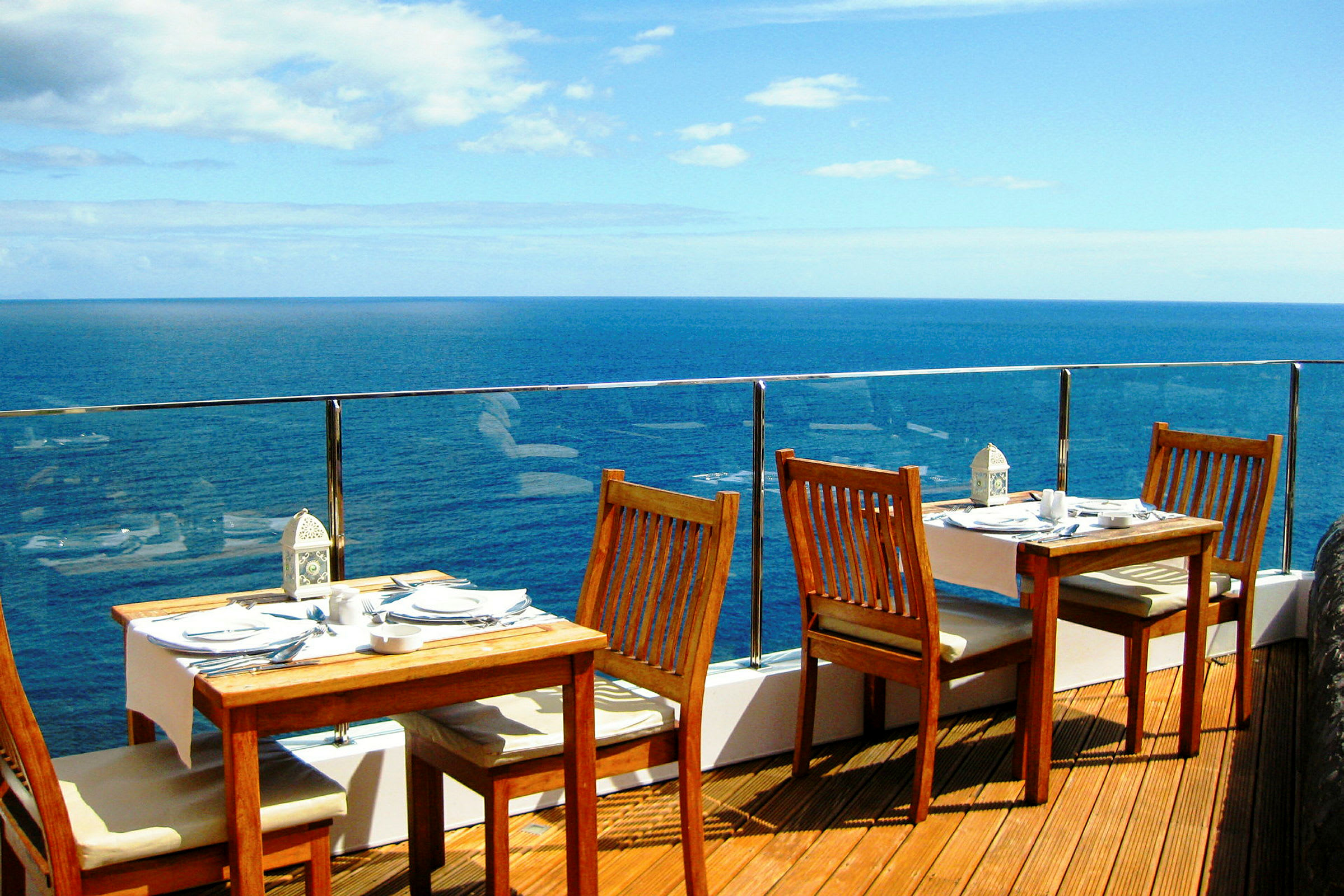Billede af en terrasse med træborde og stole ved et glasrækværk med udsigt over havet. (AI-genereret tekst)