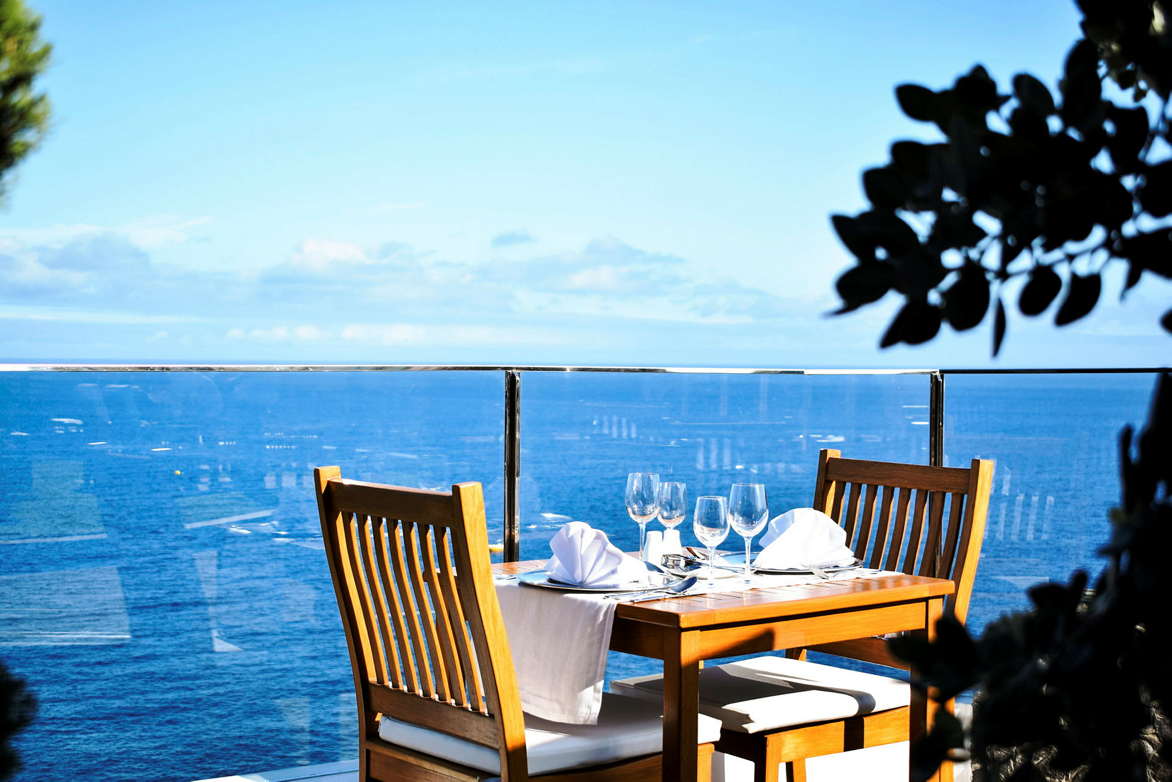 Et dækket bord med træstole og hvid dug ved et glasrækværk med udsigt over havet. (AI-genereret tekst)