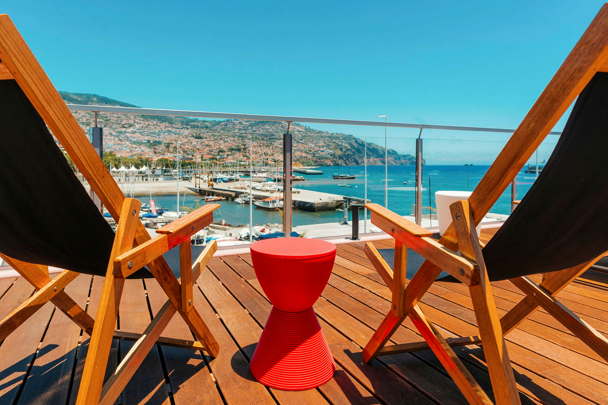 To strandstole af træ står på en terrasse med en rød lille skammel imellem. I baggrunden ses et havneområde og havet. (AI-genereret tekst)