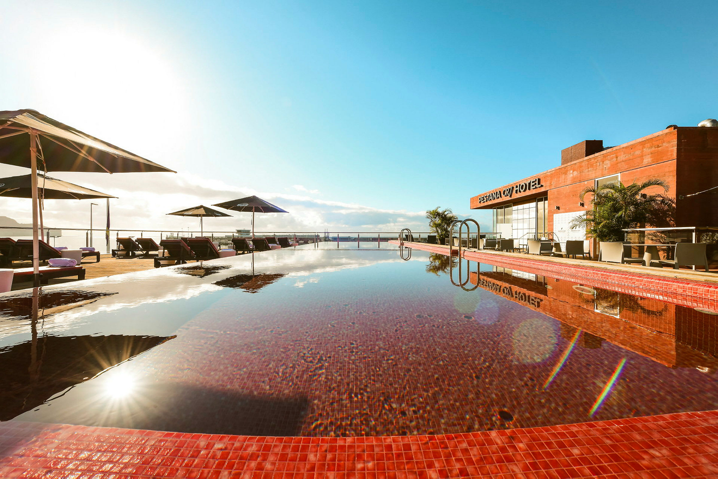 Udendørs swimmingpool med røde kanter, liggestole og parasoller under en klar himmel. (AI-genereret tekst)