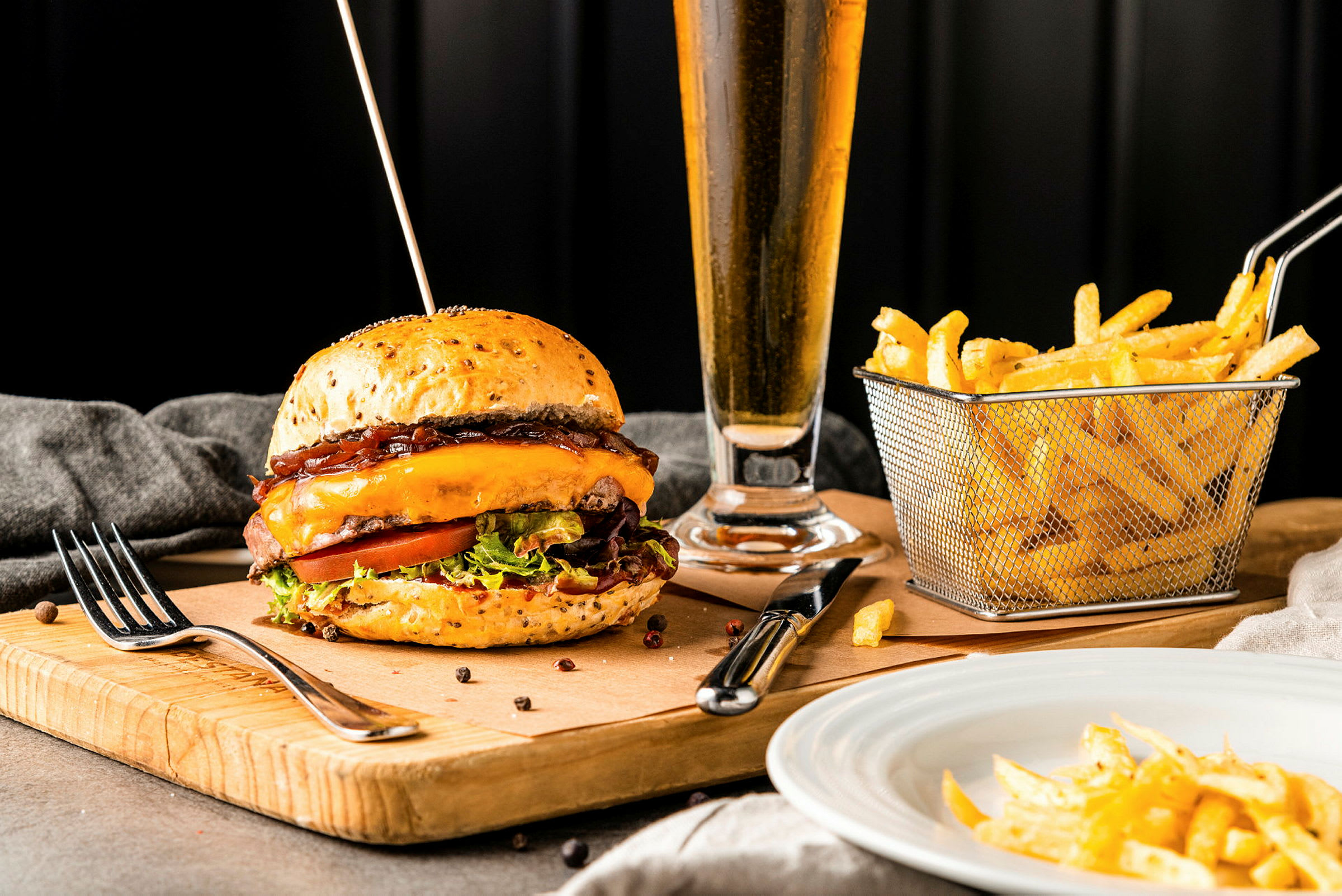 En burger med ost og salat på et træbræt, med pommes frites i en kurv og et glas drikkevarer i baggrunden. (AI-genereret tekst)