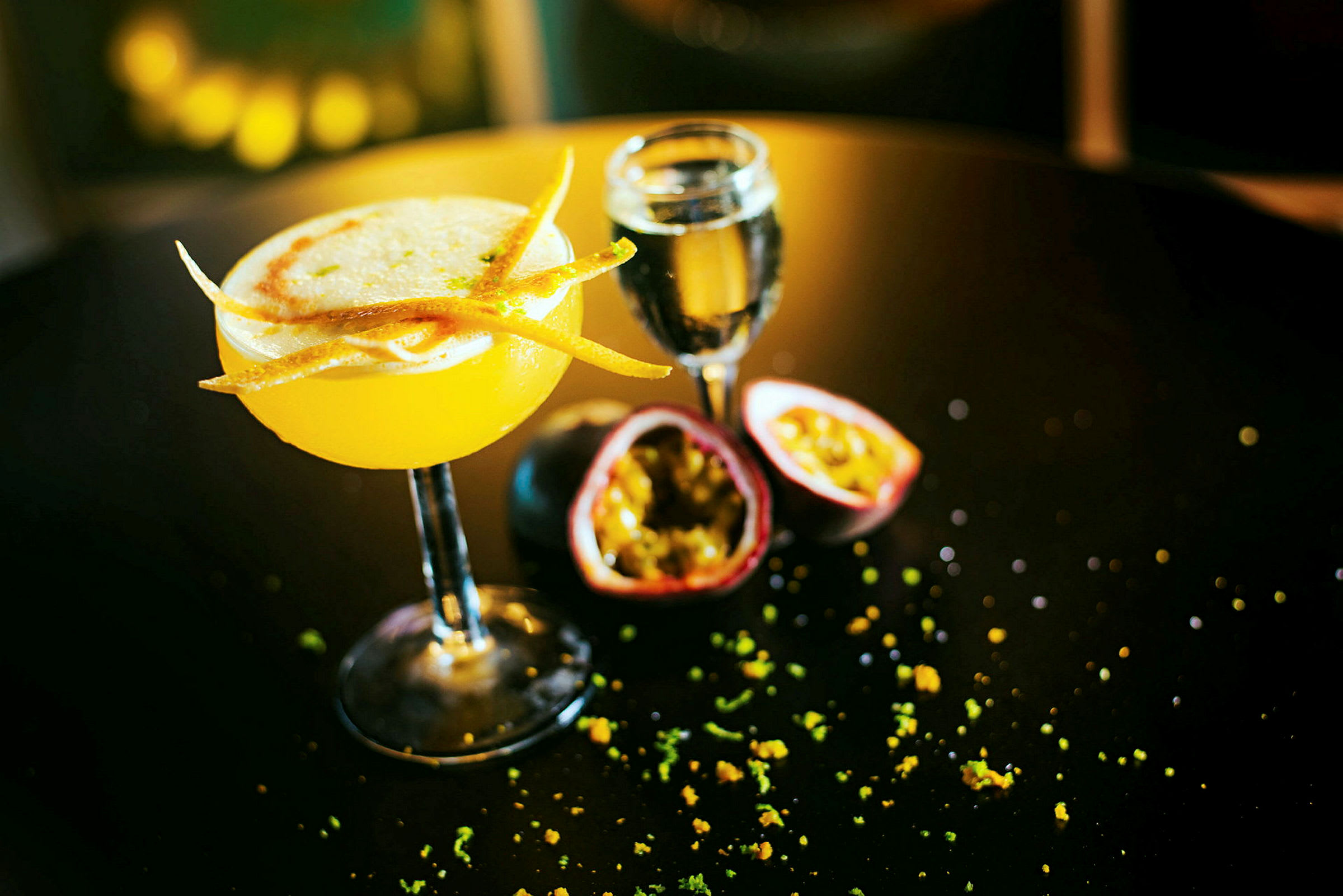 En gul cocktail med dekorationer på et bord, ved siden af et glas og halveret passionsfrugt. (AI-genereret tekst)