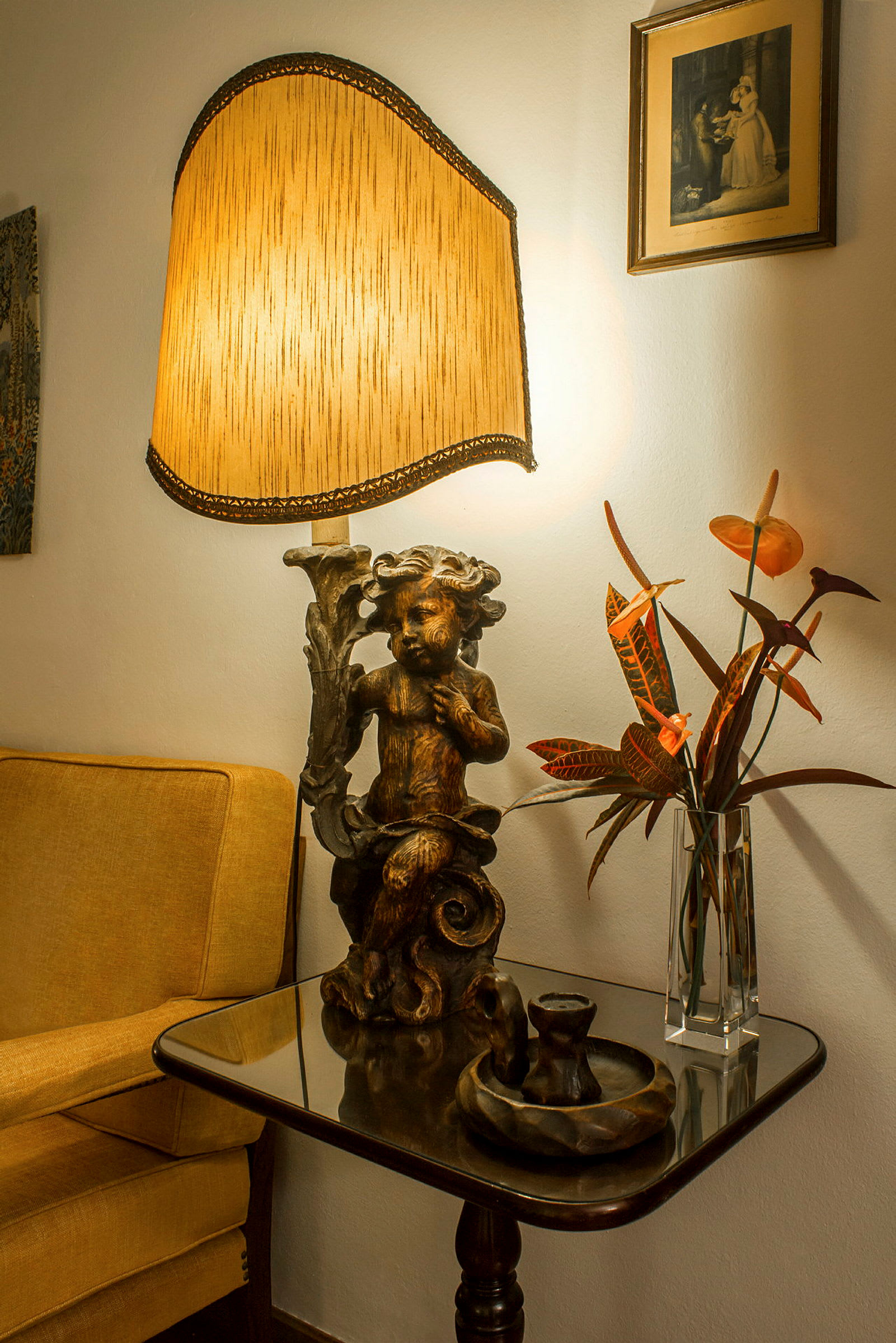Et sidebord med en dekorativ lampe, en vase med blomster og andre genstande, ved siden af en gul lænestol og et billede på væggen. (AI-genereret tekst)